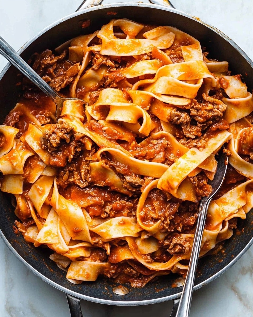 Short Ribs with Pappardelle in Rich Tomato and Red Wine Ragu, tender short ribs, hearty tomato red wine ragu, pappardelle pasta recipe, slow-cooked beef pasta dish A large pan filled with wide, flat pasta noodles mixed with shredded dark brown meat and a reddish-orange sauce coating the pasta evenly. The pasta sheets appear soft and slightly curled at the edges, layered throughout the pan with chunks of tender meat dispersed evenly in between. Two silver utensils rest on the pasta, one partially lifting some noodles, showing the noodles’ smooth and glossy texture. The pan sits on a white marbled surface.