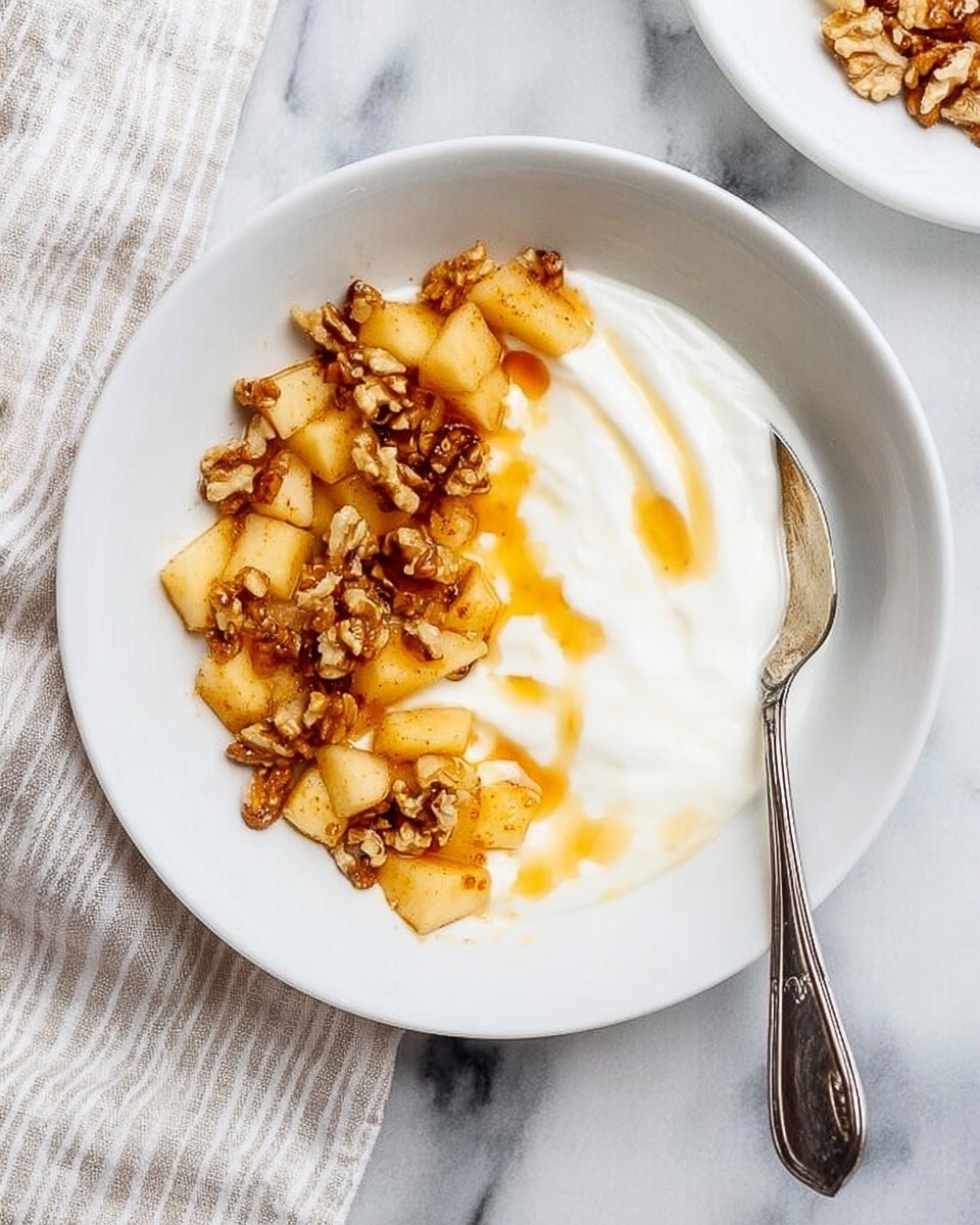 The image shows a white bowl with a smooth layer of creamy white yogurt covering about half of the bowl. On the other half, there is a topping made of small golden brown cooked apple pieces layered with chopped walnuts and a drizzle of amber honey or syrup, giving it a sticky shine. A silver spoon is placed inside the bowl, resting on the yogurt near the edge. The bowl sits on a white marbled surface with a soft striped cloth partially visible to the side. Photo taken with an iphone --ar 4:5 --v 7