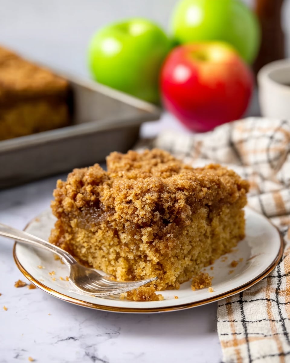 A thick square piece of brown crumb cake with a crumbly texture on top rests on a small white plate with a thin gold rim; the cake shows a moist and soft inside with small crumbs falling around it. A silver fork lies next to the cake on the plate. In the background, there is a blurred tray with more crumb cake and a green apple and red apple placed on a white marbled surface partially covered by a beige and white checkered cloth. Photo taken with an iphone --ar 4:5 --v 7