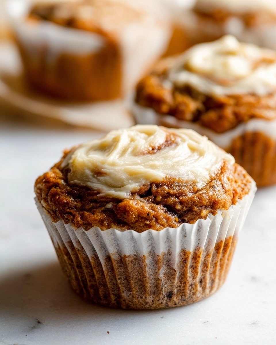 A close-up of a single muffin with a slightly rough, golden brown top with swirls of creamy white frosting spread unevenly on the surface. The muffin is nestled inside a crinkled white paper liner, which sits on a white marbled surface. The texture of the muffin looks moist and soft, with visible small darker spots in the batter. In the background, there are blurred parts of other similar muffins. Photo taken with an iphone --ar 4:5 --v 7