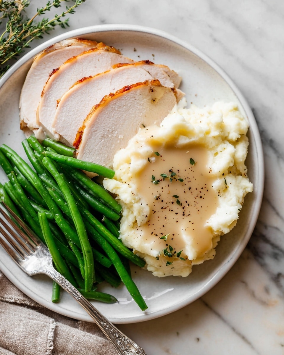 A white round plate sits on a white marbled surface holding four slices of light brown turkey arranged in a loose fan on the bottom left. Next to the turkey on the right is a mound of creamy white mashed potatoes with a smooth beige gravy pool on top, sprinkled with dark black pepper flakes and small green herbs. Above the potatoes is a neat pile of fresh bright green beans, extending horizontally across the plate. A silver fork rests with its handle extending off the bottom edge of the plate near the turkey. Photo taken with an iphone --ar 4:5 --v 7