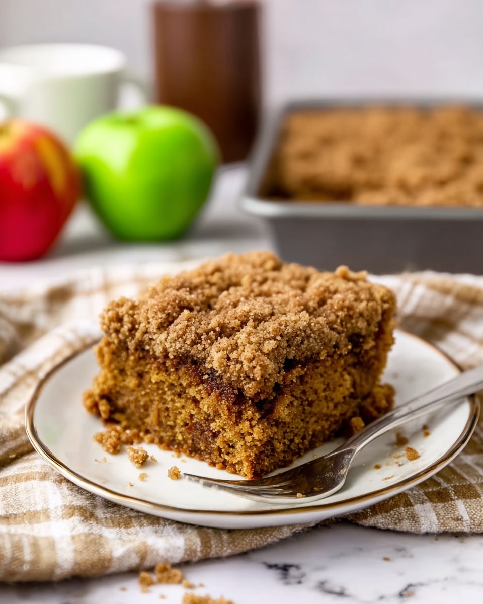 A close-up photo of a large square piece of crumbly brown cake with a rough, crumb texture on a speckled white plate. The cake shows thick, uneven layers of soft brown crumb on all sides. A woman's hand holds a metal fork pressing into the side of the cake as if about to take a bite. In the blurry background, there is a rectangular white dish filled with the same cake, and two green apples sit nearby on a white marbled surface with a folded cloth napkin. Photo taken with an iphone --ar 4:5 --v 7