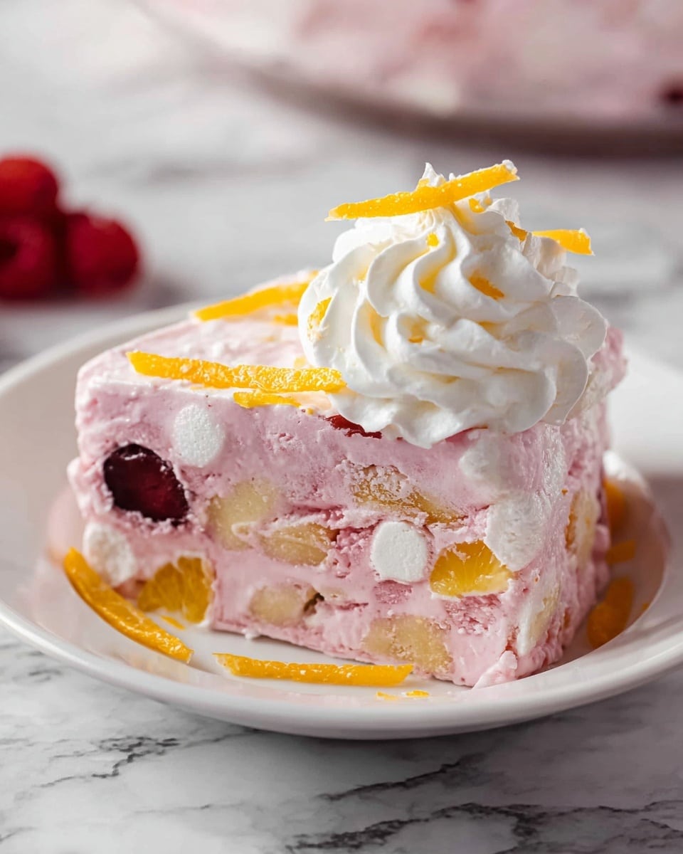 A thick slice of pink frozen dessert sits on a white plate placed on a white marbled surface. The dessert shows three visible layers of light pink creamy texture mixed with chunks of yellowish fruit and white marshmallows, plus a single dark red fruit piece in the middle. On top of the slice is a swirl of white whipped cream garnished with thin orange peel strips scattered around it. Photo taken with an iphone --ar 4:5 --v 7