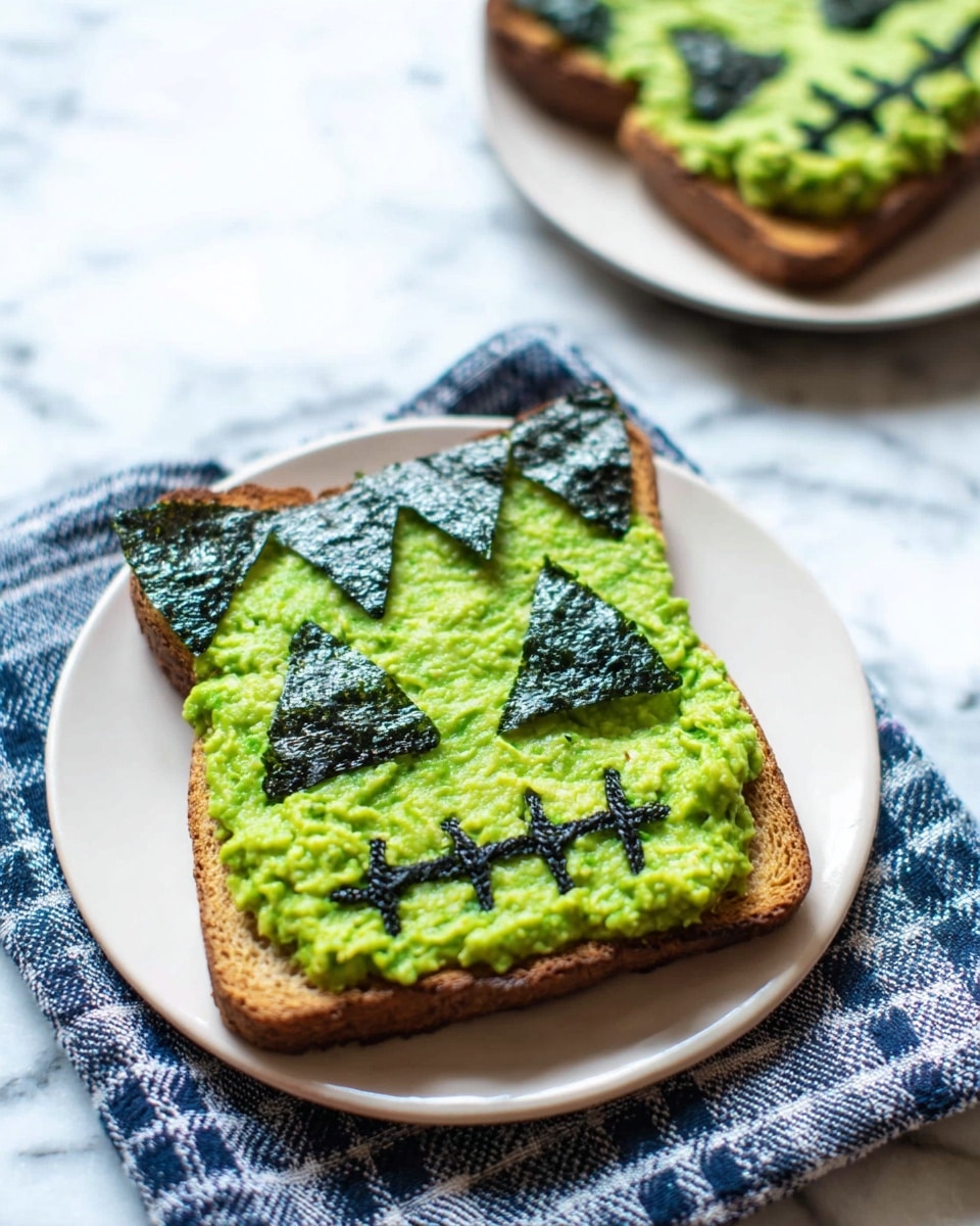 A single piece of toasted brown bread is placed on a white plate, resting on a blue and white checkered cloth over a white marbled surface. The bread is covered with a thick, uneven layer of bright green mashed avocado spread across the whole slice. On top of this, pieces of dark green seaweed are carefully arranged to form a zigzag pattern along the top edge resembling hair, two small triangles for eyes, a slim horizontal rectangle for a nose, and a stitched mouth made from connected seaweed rectangles, creating a face design. In the background, another similar toast is partially visible. photo taken with an iphone --ar 4:5 --v 7