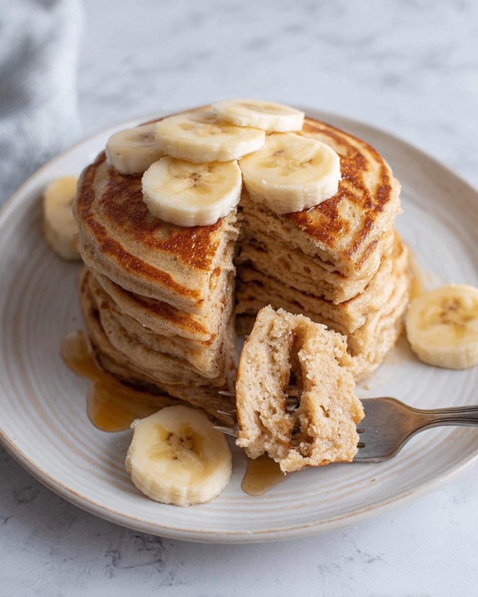 A stack of four thick, light brown pancakes with a smooth, slightly textured surface sits in the center of a white plate with a subtle raised edge design. The top pancake is decorated with three round slices of banana, which are pale yellow with soft brown accents in the center. Two more banana slices rest on the plate near the base of the stack. A fork is lifting a piece from the middle of the pancakes, showing the soft, fluffy inside with its light beige color and airy texture. A small drop of syrup is visible near the bananas on the plate. The background features a white marbled texture. Photo taken with an iphone --ar 4:5 --v 7
