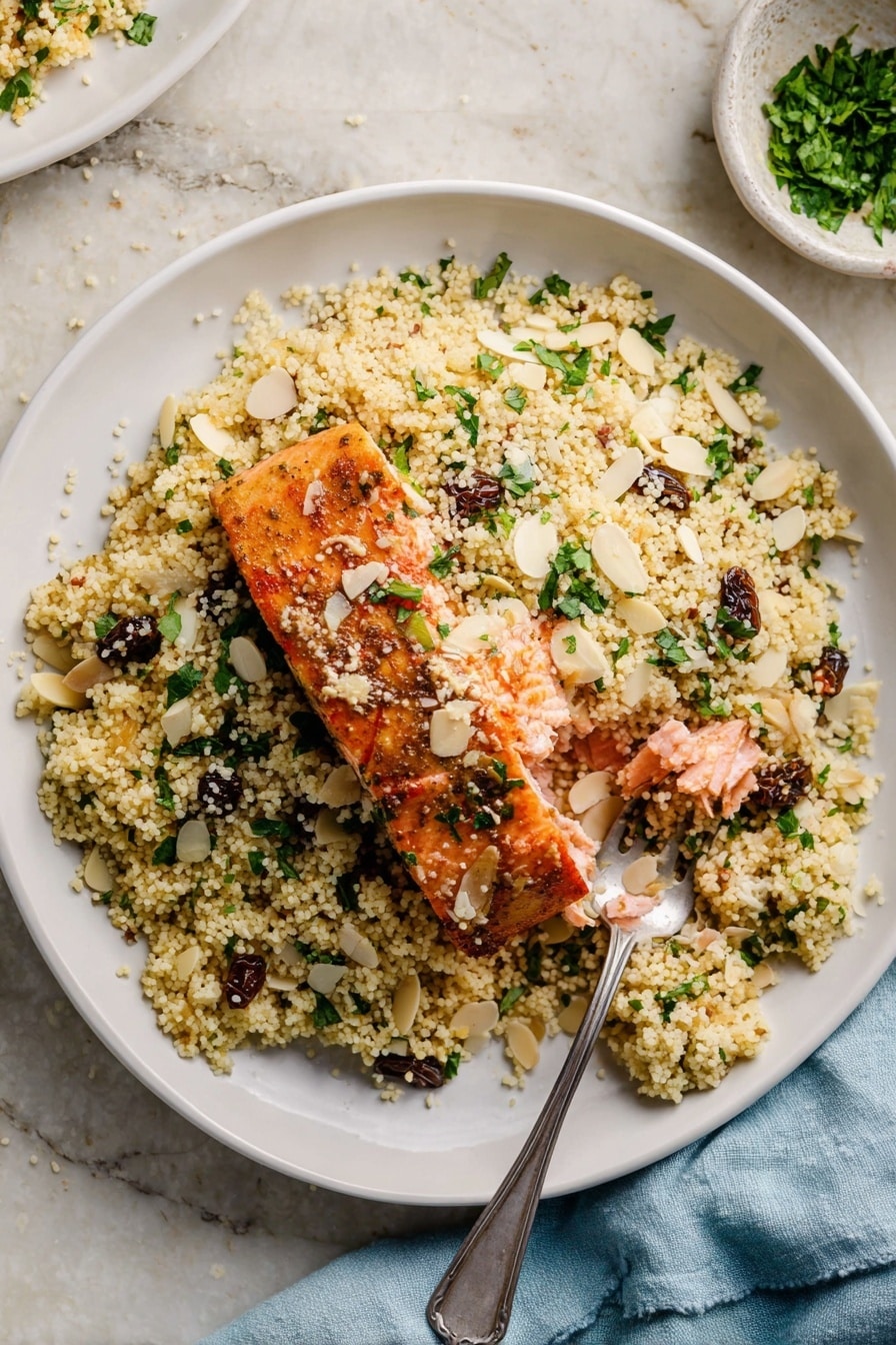 A white bowl filled with a base layer of light yellow couscous mixed with chopped green herbs and small dark raisins. On top of the couscous, there is a piece of cooked salmon with a golden brown crust, broken open to show the soft pink inside. Thin almond slices and extra chopped green herbs are scattered on the dish. A silver spoon rests on the right side of the bowl, partially under the couscous. The bowl sits on a white marbled surface, and a light blue cloth napkin is visible nearby. photo taken with an iphone --ar 2:3 --v 7 - Moroccan-Spiced Salmon and Couscous, Moroccan-Spiced Salmon, Couscous Recipes, Quick Fish Dinner, Easy Weeknight Dinner
