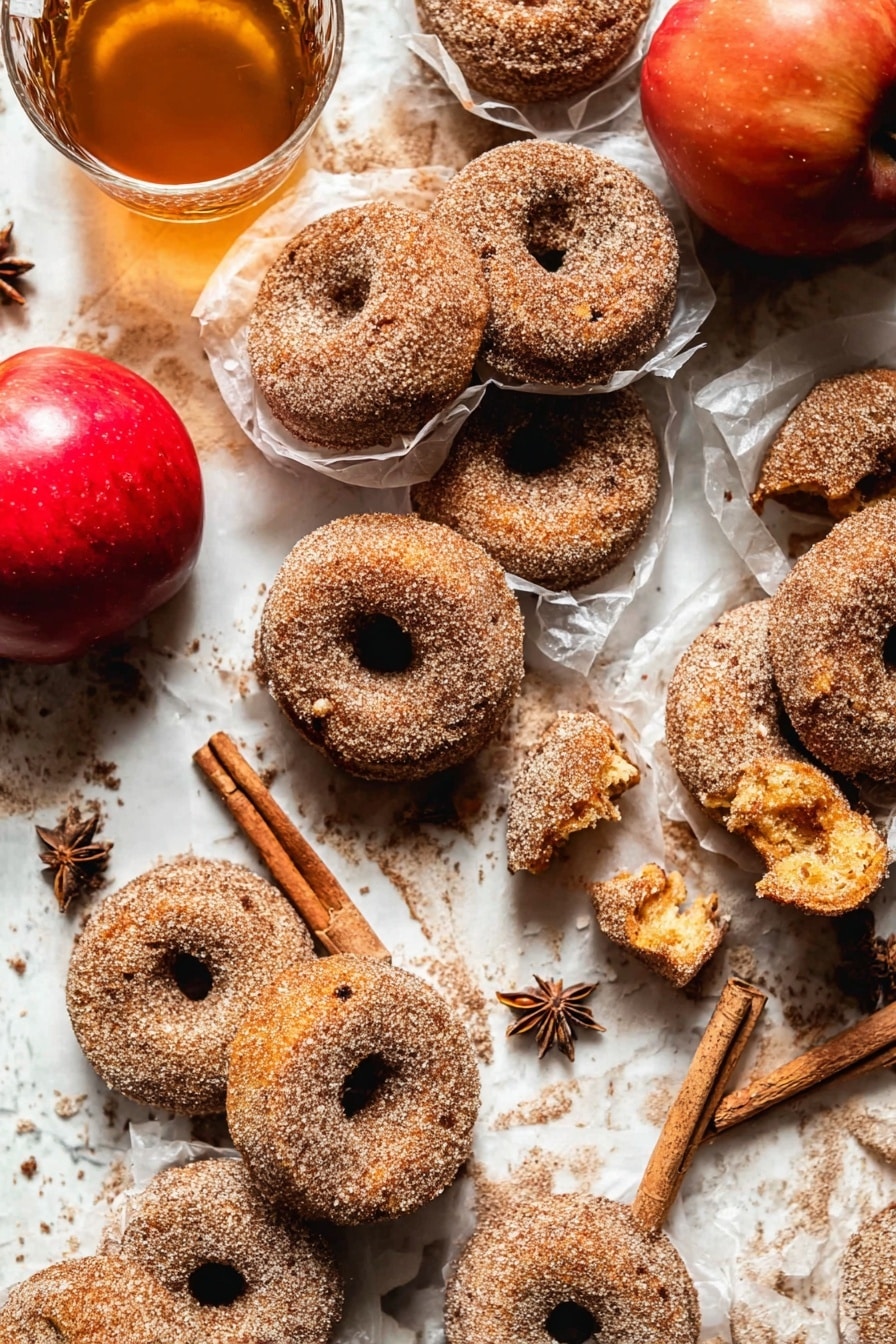 The image shows many cinnamon sugar donuts placed closely on a white marbled surface. Most donuts are whole, with a rough texture covered in sugar and cinnamon. A few donuts are broken, revealing a soft, golden inside with a slightly crumbly texture. Some donuts are wrapped in thin, translucent white paper, adding layers and depth. Near the donuts, there is a bright red apple and a clear glass cup filled with light brown tea and cinnamon sticks inside. Star anise is scattered around for decoration. The scene looks warm and cozy with soft natural light. Photo taken with an iphone --ar 2:3 --v 7 - Baked Cinnamon Crunch Apple Cider Doughnuts, autumn apple cider donuts, baked fall doughnuts, cinnamon sugar baked doughnuts, no-fry apple cider treats