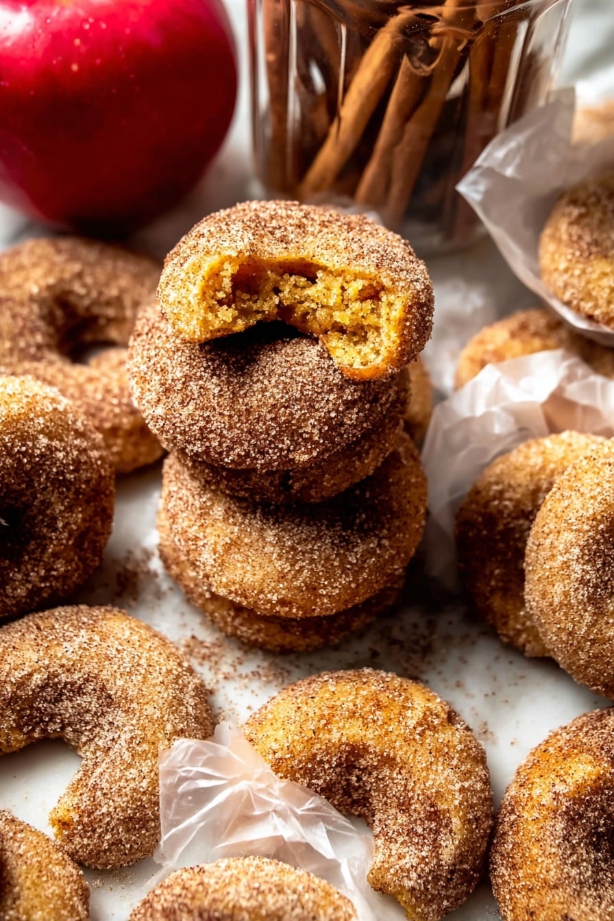 The image shows several crescent-shaped cookies covered in a coarse layer of cinnamon sugar. The cookies are golden brown with a slightly rough texture beneath the sugar coating. Some cookies are stacked in a loose pile, while others are individually wrapped in translucent white paper. The cookies have a crumbly look, with one cookie broken open to reveal a soft yellow interior. In the background, there is a bright red apple and a clear glass jar filled with more cookies and cinnamon sticks on top. The setting uses a white marbled surface underneath all the items. photo taken with an iphone --ar 2:3 --v 7 - Baked Cinnamon Crunch Apple Cider Doughnuts, autumn apple cider donuts, baked fall doughnuts, cinnamon sugar baked doughnuts, no-fry apple cider treats