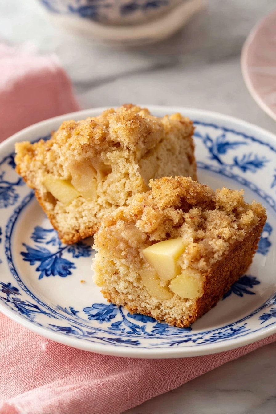 Two pieces of crumb-topped cake are placed on a white plate with blue floral patterns. The cake has a light brown color with soft, crumbly texture and visible chunks of yellow apple inside. The crumb topping is thicker and golden brown. The plate sits on a pink cloth, all set on a white marbled surface. Photo taken with an iphone --ar 2:3 --v 7 - Apple Muffins Recipe, moist apple muffins, cinnamon apple muffins, easy apple muffin recipe, homemade apple muffins