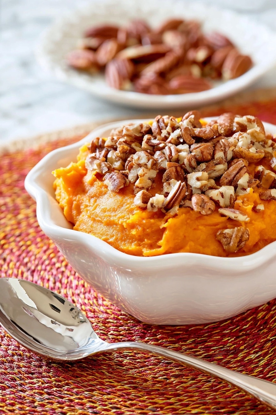 This image shows a white bowl filled with a thick, mashed orange sweet potato layer that is roughly shaped with a soft texture. On top of this layer, there is a generous amount of chopped pecans scattered unevenly, adding a crunchy contrast with their brown, nutty pieces. The bowl sits on a woven orange, red, and yellow circular placemat, and in the blurred background, part of another white dish with light brown pecans can be seen resting on a white marbled surface. photo taken with an iphone --ar 2:3 --v 7 - Slow Cooker Sweet Potato Casserole with Toasted Pecans, sweet potato casserole, slow cooker sweet potato side dish, holiday sweet potato recipes, easy slow cooker side dish