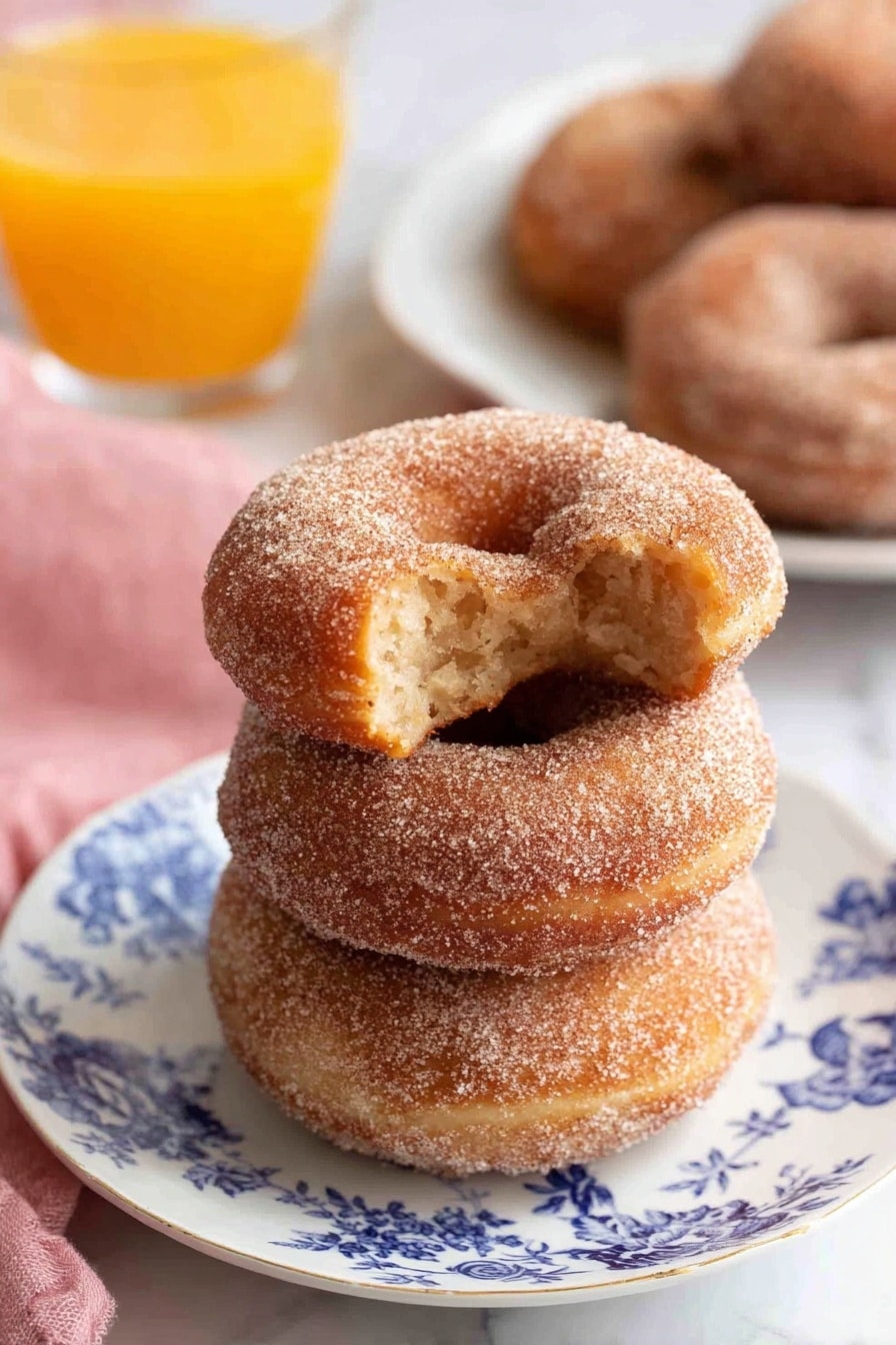 Three cinnamon sugar donuts are stacked on a white plate with blue floral designs, showing a soft texture covered in fine sugar crystals; the top donut has a bite taken, revealing a light, fluffy inside with a slightly darker core. Behind the plate is a blurred white plate with more cinnamon sugar donuts and a clear glass filled with bright orange juice, all set on a white marbled surface. A pink cloth is placed softly on the left side. photo taken with an iphone --ar 2:3 --v 7 - Apple Cider Donuts, Apple Cider Donuts Recipe, Fall Donuts, Homemade Apple Cider Donuts, Spiced Donut Recipe