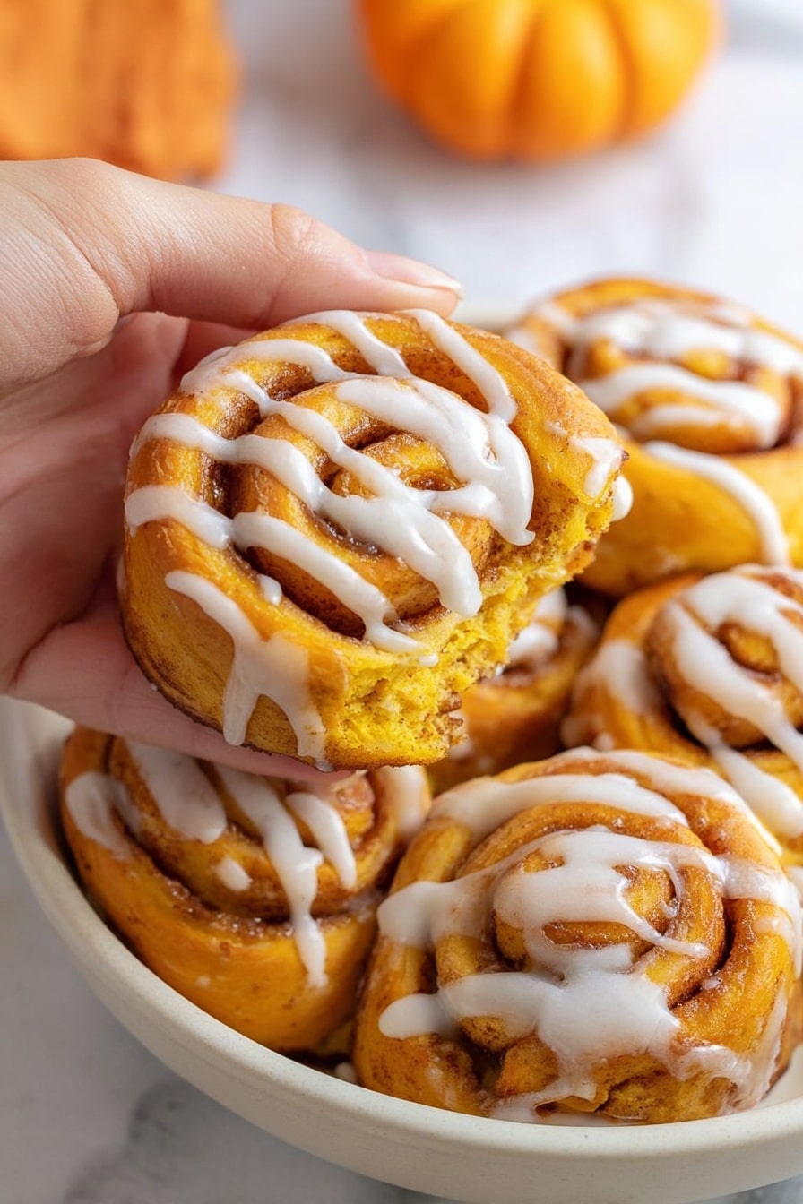 A woman's hand is holding one bright orange swirl roll with white icing drizzled across the surface, showing its soft and fluffy texture with cinnamon filling layers. Around it are several more identical rolls placed close together in a white bowl, all with the same orange color and white icing in spiral patterns. The background shows a white marbled surface with part of a small orange pumpkin visible on the right side. The overall scene highlights the warm, rich colors and creamy icing on the rolls. photo taken with an iphone --ar 2:3 --v 7 - Pumpkin Cinnamon Rolls, easy fall breakfast, no yeast cinnamon rolls, pumpkin dessert recipes, quick pumpkin sweet rolls