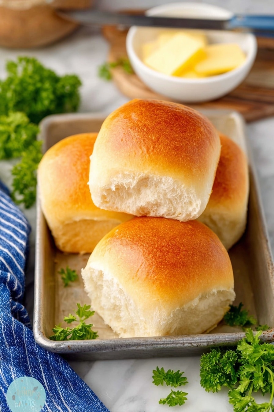 The image shows four soft, shiny bread rolls with a golden brown top, stacked and placed inside a metal baking tray on a white marbled surface. The bread looks fluffy and light, with a slightly textured white inside. Behind the rolls, there is a small white bowl holding a pale yellow stick of butter and a butter knife resting on the bowl's edge. Bright green parsley leaves are scattered around the tray, adding a fresh touch. A blue and white striped cloth is partly visible on the left side, contributing to a cozy, homey feel. photo taken with an iphone --ar 2:3 --v 7 - Fluffy Homemade Dinner Rolls, Soft dinner roll recipe, buttery dinner rolls, easy homemade bread, bakery-style dinner rolls