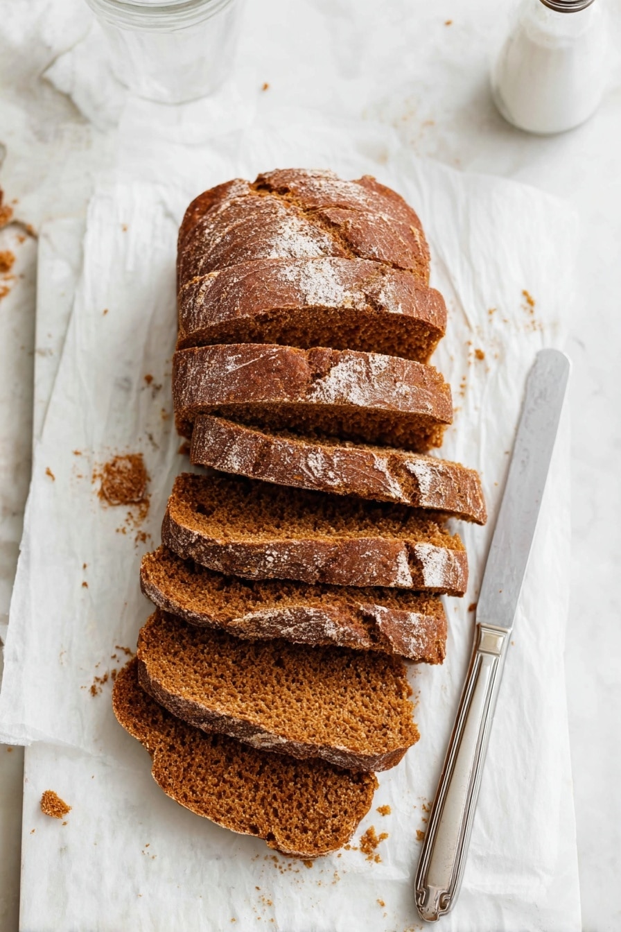The image shows a loaf of brown bread cut into six thick slices, placed slightly overlapping each other on a white parchment paper. The bread has a textured crust with a few light flour spots on top and a soft, crumbly inside with a warm brown color. To the right of the bread, there is a silver knife with a simple handle resting on the parchment paper. The background is a white marbled surface with some bread crumbs scattered around, and the top right part of a clear glass and a white salt shaker are visible. Photo taken with an iphone --ar 2:3 --v 7 - Moist Pumpkin Bread, pumpkin bread recipe, easy pumpkin bread, autumn pumpkin bread, cozy pumpkin bread