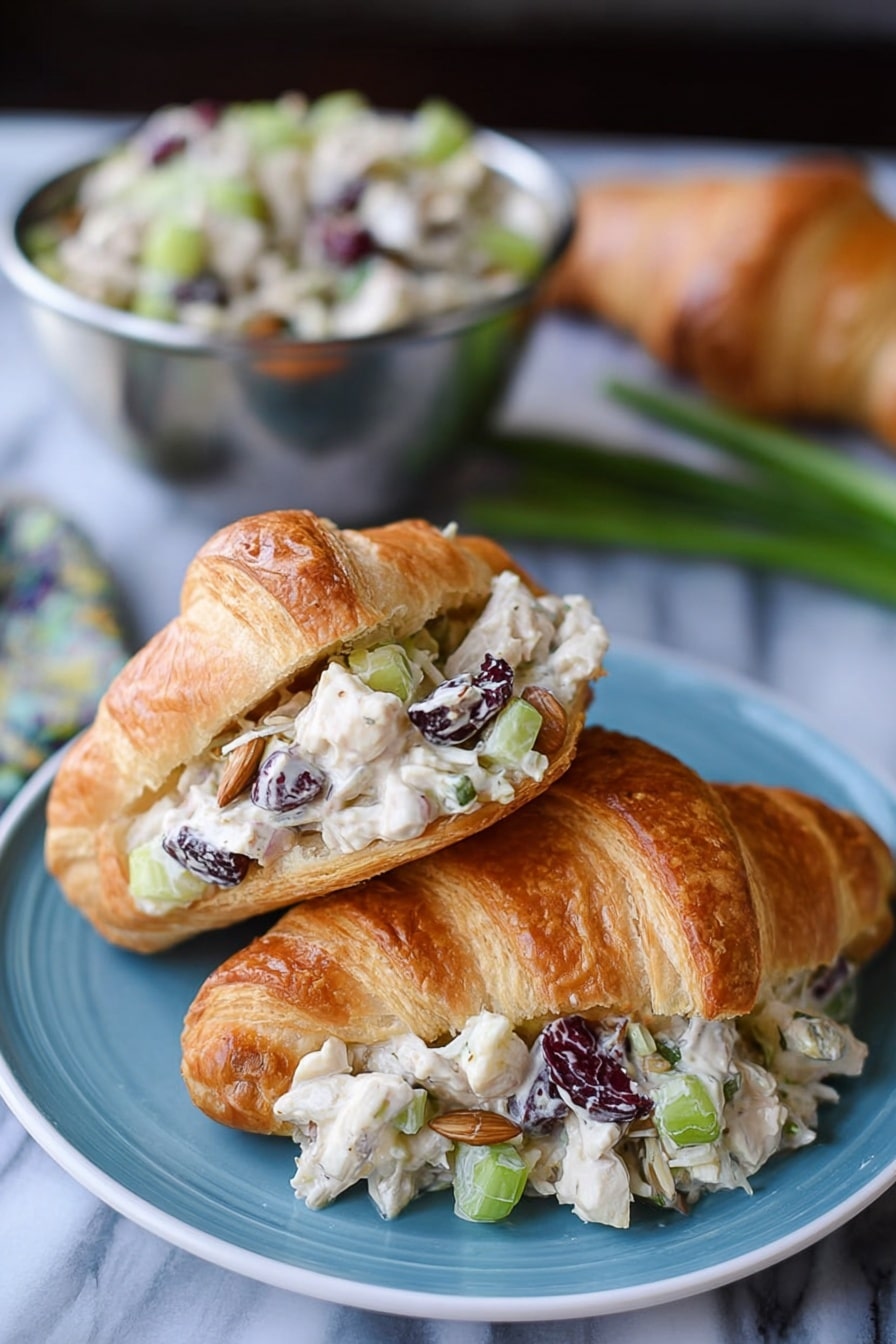 The image shows two croissants on a blue plate placed on a white marbled surface. One croissant is whole with a golden-brown, flaky texture. The other croissant is open and filled with a creamy salad made of small white chicken pieces, chopped celery, dried dark cranberries, sunflower seeds, and light beige dressing, layered inside the croissant’s light and flaky interior. In the background, there is a metal bowl filled with more of the salad, and some green onions and an additional croissant are blurred. Photo taken with an iphone --ar 2:3 --v 7 - Cranberry Chicken Salad, Cranberry Chicken Salad Recipe, Easy Chicken Salad with Cranberries, Quick Cranberry Chicken Salad, Festive Chicken Salad