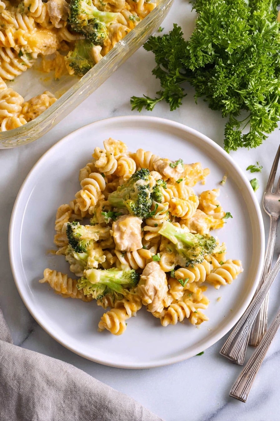 A white plate holds a serving of pasta made with spiral-shaped noodles mixed with chunks of light-colored cooked chicken and small, bright green broccoli florets, all coated in a creamy light yellow cheese sauce with a slightly shiny texture; the dish is placed on a white marbled surface next to a partially visible glass baking dish filled with more pasta, a small bunch of fresh parsley with deep green leaves, and a folded light gray cloth napkin holding three metal forks. Photo taken with an iphone --ar 2:3 --v 7 - Creamy Chicken Broccoli Noodle Casserole, cheesy chicken broccoli bake, easy comfort food dinner, hearty chicken noodle casserole, quick weeknight family meal