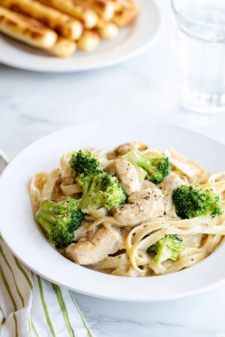 A white plate holds a serving of creamy fettuccine pasta mixed with light beige chicken pieces and bright green broccoli florets, all coated in a smooth, pale sauce with a few black pepper specks on top. In the background, out of focus, there is a white plate with golden brown breadsticks and a glass of water next to it. The scene is set on a white marbled surface with a folded white and green striped cloth visible beside the plate. photo taken with an iphone --ar 2:3 --v 7 - Creamy Chicken Broccoli Fettuccine Alfredo, Chicken Fettuccine Alfredo, Easy Chicken Pasta, Homemade Alfredo Sauce, Quick Weeknight Dinner
