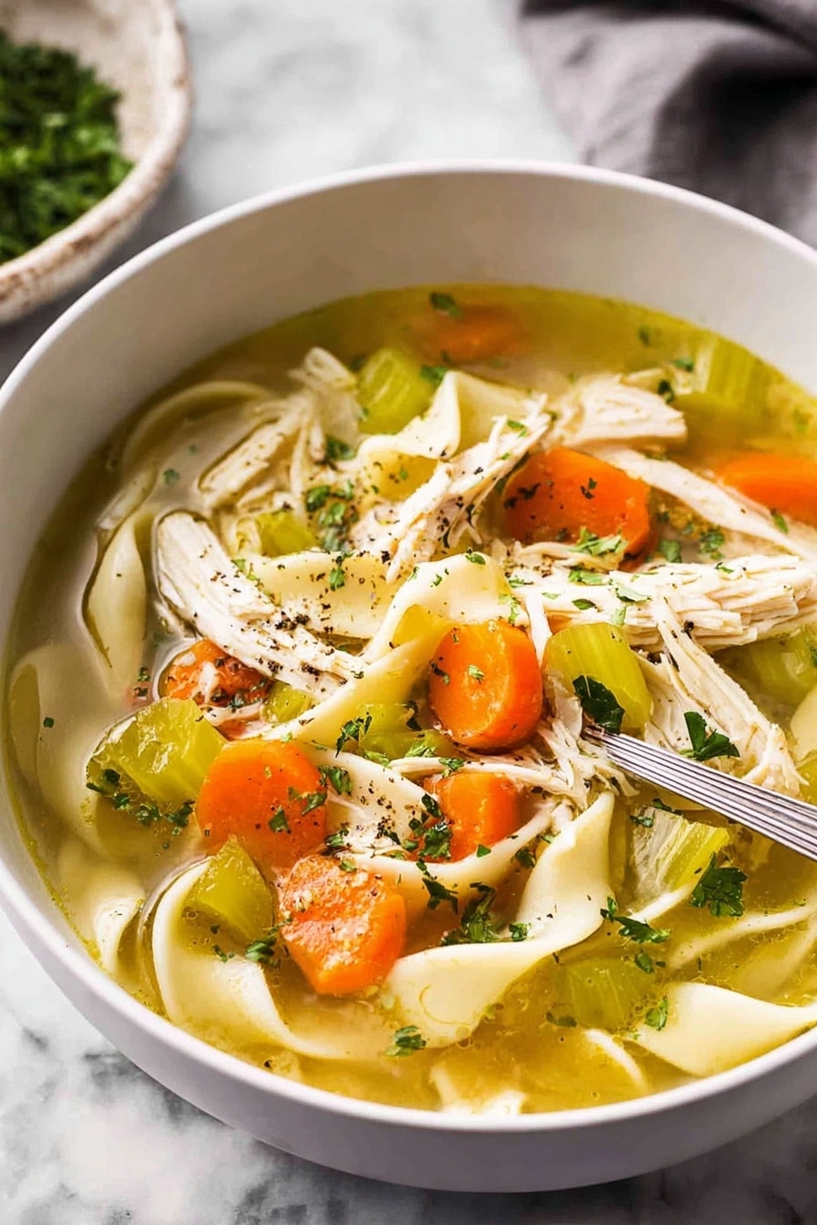 The image shows a close-up view of a white bowl filled with clear chicken soup. Inside the soup, there are wide flat noodles curled softly, bright orange carrot slices, pieces of pale green celery, and shredded white chicken meat scattered on one side. Small green parsley flakes float on the surface along with some black pepper specks. The soup is light golden and clear, with visible layers of ingredients resting naturally in the broth. The bowl sits on a white marbled surface. photo taken with an iphone --ar 2:3 --v 7 - Homemade Chicken Noodle Soup from Scratch, chicken noodle soup, homemade chicken soup, comforting chicken noodle recipe, fresh chicken soup