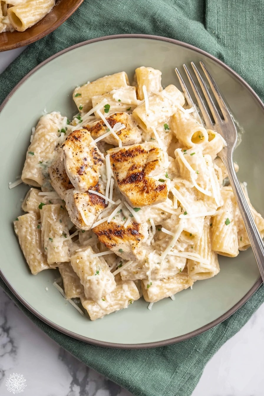 A white plate holds a serving of rigatoni pasta coated in a creamy white sauce with small green herb flecks. On top of the pasta lie four grilled chicken pieces, light brown with darker grill marks, partially covered by the sauce. Thin white shredded cheese is sprinkled over the chicken and pasta, adding texture. A silver fork rests on the right side of the plate. The plate is on a soft green cloth placed on a white marbled surface. Photo taken with an iphone --ar 2:3 --v 7 - One Pot Creamy Parmesan Chicken Pasta, simple chicken pasta dish, easy one pot dinner, cheesy chicken pasta recipe, weeknight chicken pasta