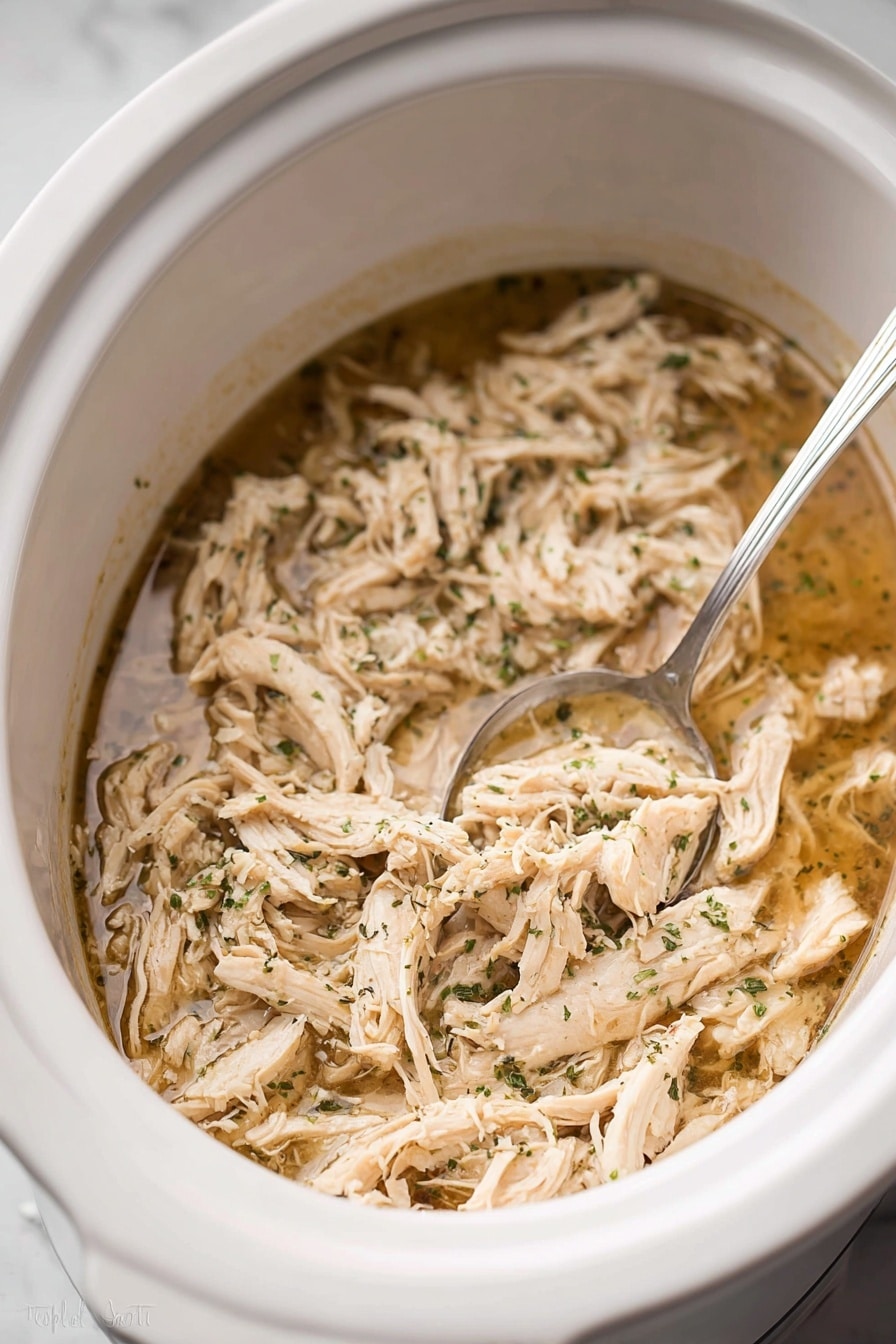 Inside a white slow cooker, there is one main layer of shredded light beige cooked chicken mixed with small green herb pieces, all soaked in a thin, light brown broth. A silver spoon rests partially submerged in the mixture on the right side. The chicken strands have a soft texture, and the herbs are finely sprinkled. The slow cooker sits on a white marbled surface. photo taken with an iphone --ar 2:3 --v 7 - Juicy Crockpot Shredded Chicken, Crockpot shredded chicken recipe, easy slow cooker chicken, tender shredded chicken, versatile chicken recipes