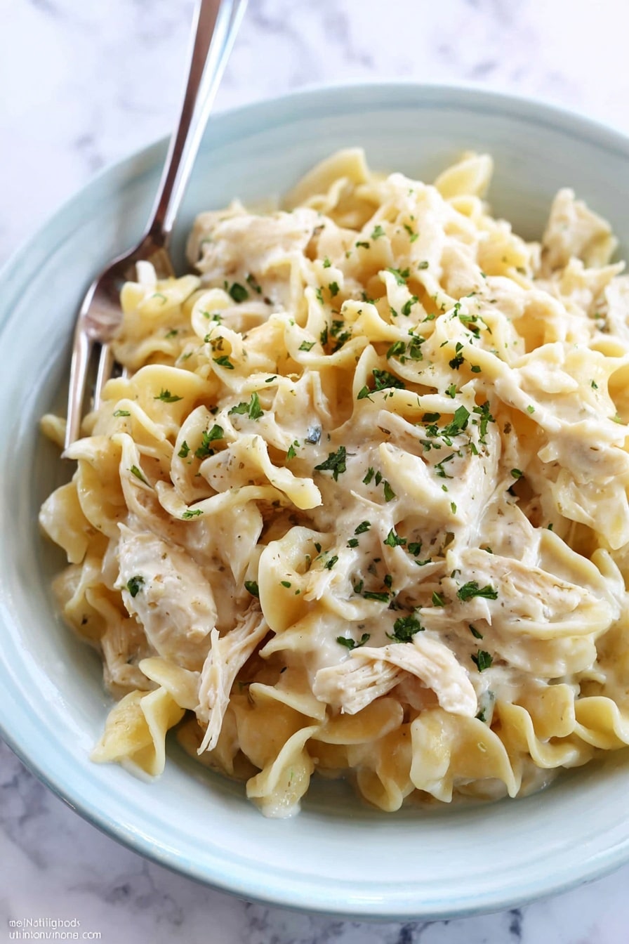 A close-up of creamy chicken noodle dish served in a white bowl placed on a white marbled surface. The dish has three main layers: at the bottom is a thick white cream sauce, on top of it a layer of soft cooked egg noodles with a slightly wavy texture and pale yellow color, and scattered throughout and on top are pieces of shredded chicken mixed well with the noodles. The whole dish is sprinkled with small green herb flakes for garnish. A fork is resting inside the bowl, ready to eat. photo taken with an iphone --ar 2:3 --v 7 - Crockpot Chicken and Noodles, slow cooker chicken pasta, easy chicken noodle meal, comforting crockpot dinner, creamy chicken and noodles