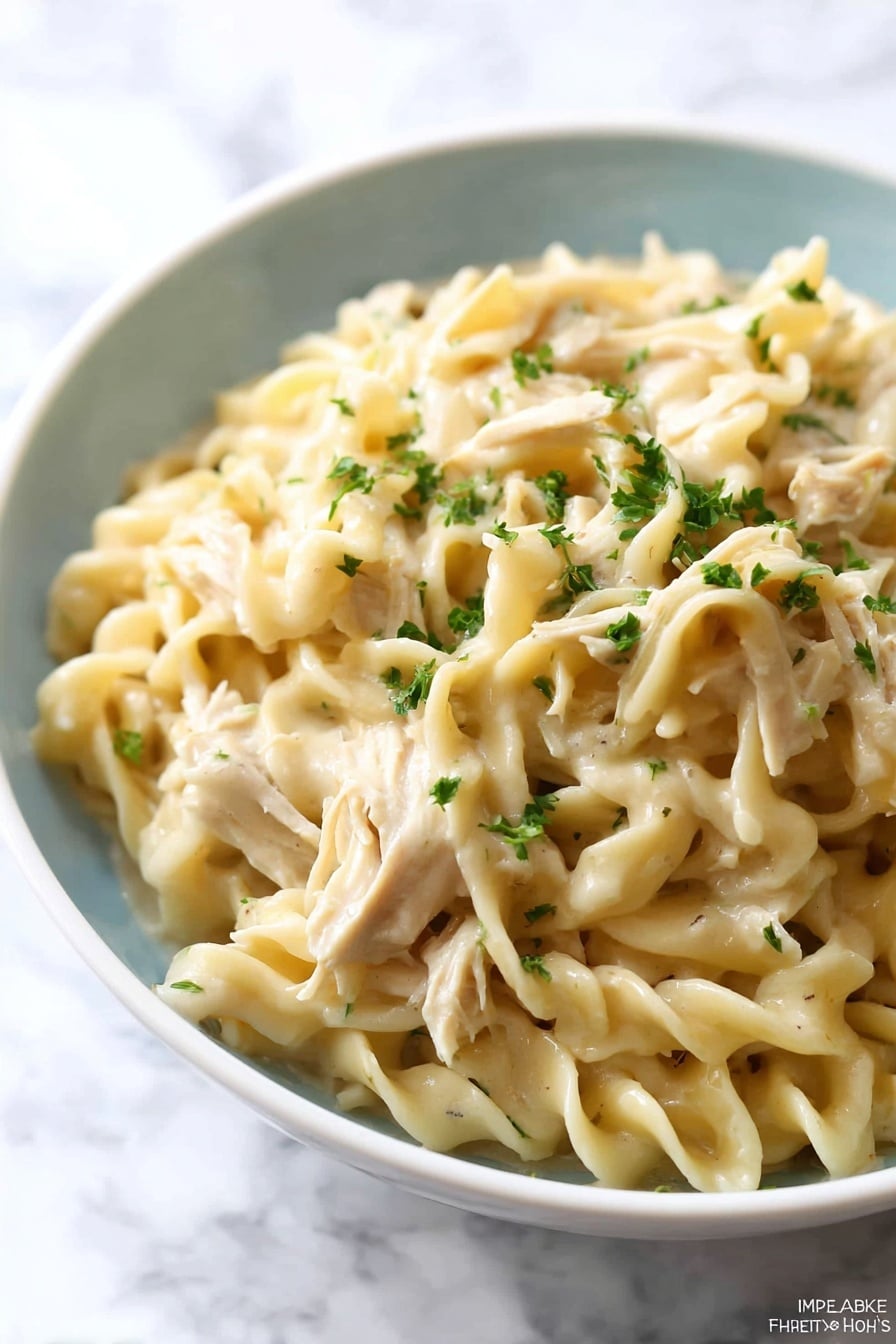 A close-up of a white bowl filled with creamy egg noodles mixed with shredded pieces of light beige chicken. The egg noodles are thick and twisted, coated in a smooth, light cream sauce. Small bits of green herbs are sprinkled on top and some are mixed within the noodles, adding small dots of green color. The bowl is set on a white marbled surface, creating a clean and bright background. Photo taken with an iphone --ar 2:3 --v 7 - Crockpot Chicken and Noodles, slow cooker chicken pasta, easy chicken noodle meal, comforting crockpot dinner, creamy chicken and noodles