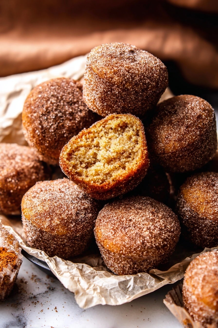 The image shows a pile of small, round doughnut muffins covered in cinnamon sugar on a white marbled surface. There are about ten muffins closely stacked, with one muffin on top showing its inside layer which is soft and light brown with a slightly crumbly texture. The muffins have a rough, grainy outer layer from the sugar coating, and they are resting on crumpled white parchment paper. The scene captures a warm, cozy feeling from the brown and golden hues of the muffins, with some muffins sitting inside a black tray. photo taken with an iphone --ar 2:3 --v 7 - Baked Cinnamon Sugar Apple Cider Doughnut Holes, fall apple cider treats, baked doughnut holes with cinnamon, cozy autumn snack, easy baked doughnut recipe