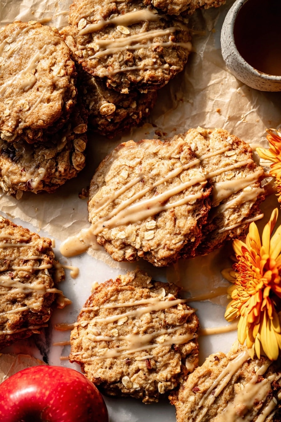 Several oatmeal cookies are spread on light brown parchment paper over a white marbled surface. The cookies are golden brown with a rough texture, showing visible oats and slightly darker edges that look crispy. Each cookie is drizzled with a thin layer of light caramel-colored glaze, creating irregular lines and patches across their tops. In the bottom left corner of the image, two whole red apples with light yellow patches are placed together, adding a fresh contrast to the warm cookie colors. The light source creates soft shadows and highlights, enhancing the cookies’ textures and the glaze's shine. photo taken with an iphone --ar 2:3 --v 7 - Iced Apple Cider Caramel Oatmeal Cookies, apple cider oatmeal cookies, caramel oatmeal cookies, fall cookies with apple cider, chewy caramel oatmeal treats