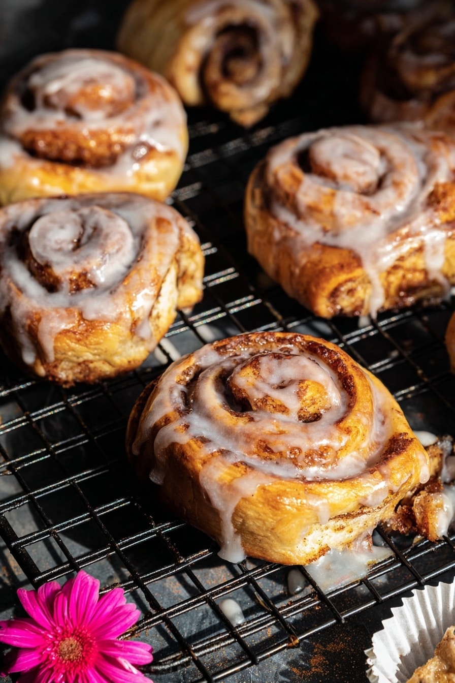 The image shows several cinnamon rolls on a black wire cooling rack with a dark base underneath. Each cinnamon roll has visible spiral layers of golden-brown dough swirled with darker brown cinnamon sugar. The top layers are coated with a shiny white glaze that drips slightly down the sides and onto the rack. The rolls have a soft, fluffy texture with a mix of light yellow and brown tones, and some darker, slightly crisped edges. Near the bottom right, there is a white paper liner with a bright pink flower placed on it. The light creates highlights on the glossy glaze and shadows between the cinnamon rolls, giving the scene depth and warmth. Photo taken with an iphone --ar 2:3 --v 7 - Shortcut Easy Apple Fritters, apple fritters with puff pastry, quick apple fritters recipe, cinnamon apple fritters, fall dessert recipes