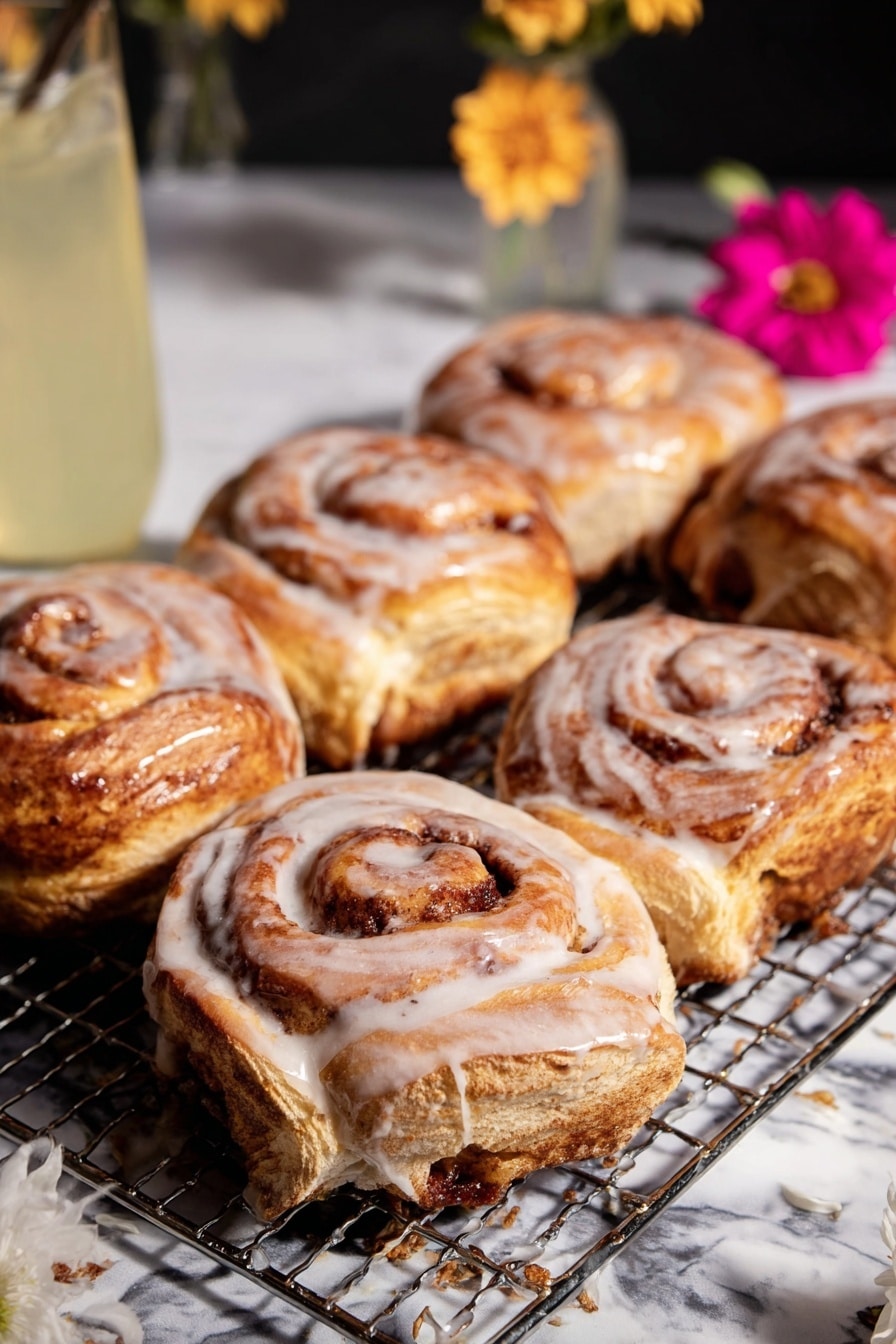 The image shows six cinnamon rolls with a thick white glaze, placed on a wire cooling rack over a white marbled surface. Each cinnamon roll has multiple golden brown layers swirled tightly together with visible cinnamon filling, some layers darker due to caramelized sugar. The glaze drips slightly down the sides, adding a shiny texture on top of the soft, fluffy layers. A white paper liner holds one cinnamon roll in the front, surrounded by a few splashes of glaze on the surface. In the background, a tall glass with a light yellow liquid and a flower in a small white vase add a soft decorative touch, along with a pink flower in a white paper liner near the lower right corner. photo taken with an iphone --ar 2:3 --v 7 - Shortcut Easy Apple Fritters, apple fritters with puff pastry, quick apple fritters recipe, cinnamon apple fritters, fall dessert recipes