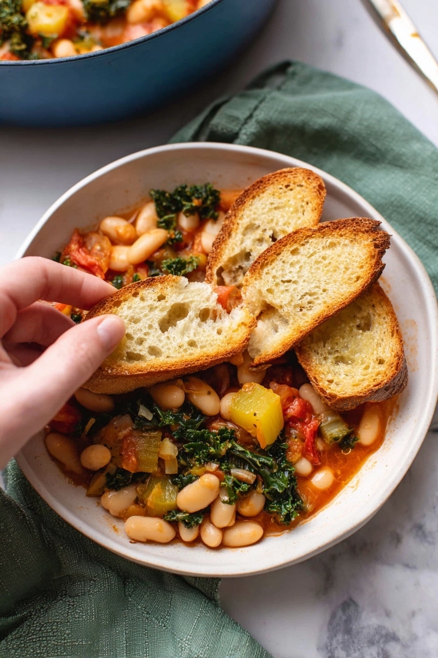 A white bowl filled with a warm stew made of white beans, chunks of yellow squash, wilted green kale, and small red tomato pieces. On top, three slices of toasted baguette bread rest, one slice held by a woman's hand, showing the mix of beans and vegetables piled on it. The stew's sauce is a light red color, and the bread has a golden-brown crust with a porous, soft inside texture. The bowl sits on a green cloth on a white marbled surface, with part of a blue pot filled with more stew blurred in the background. Photo taken with an iphone --ar 2:3 --v 7 - Stewed Great Northern Beans with Kale Tomatoes and Herb-Infused Broth, hearty bean and kale stew, healthy vegetarian bean recipe, nourishing winter dinner idea, easy bean and greens recipe