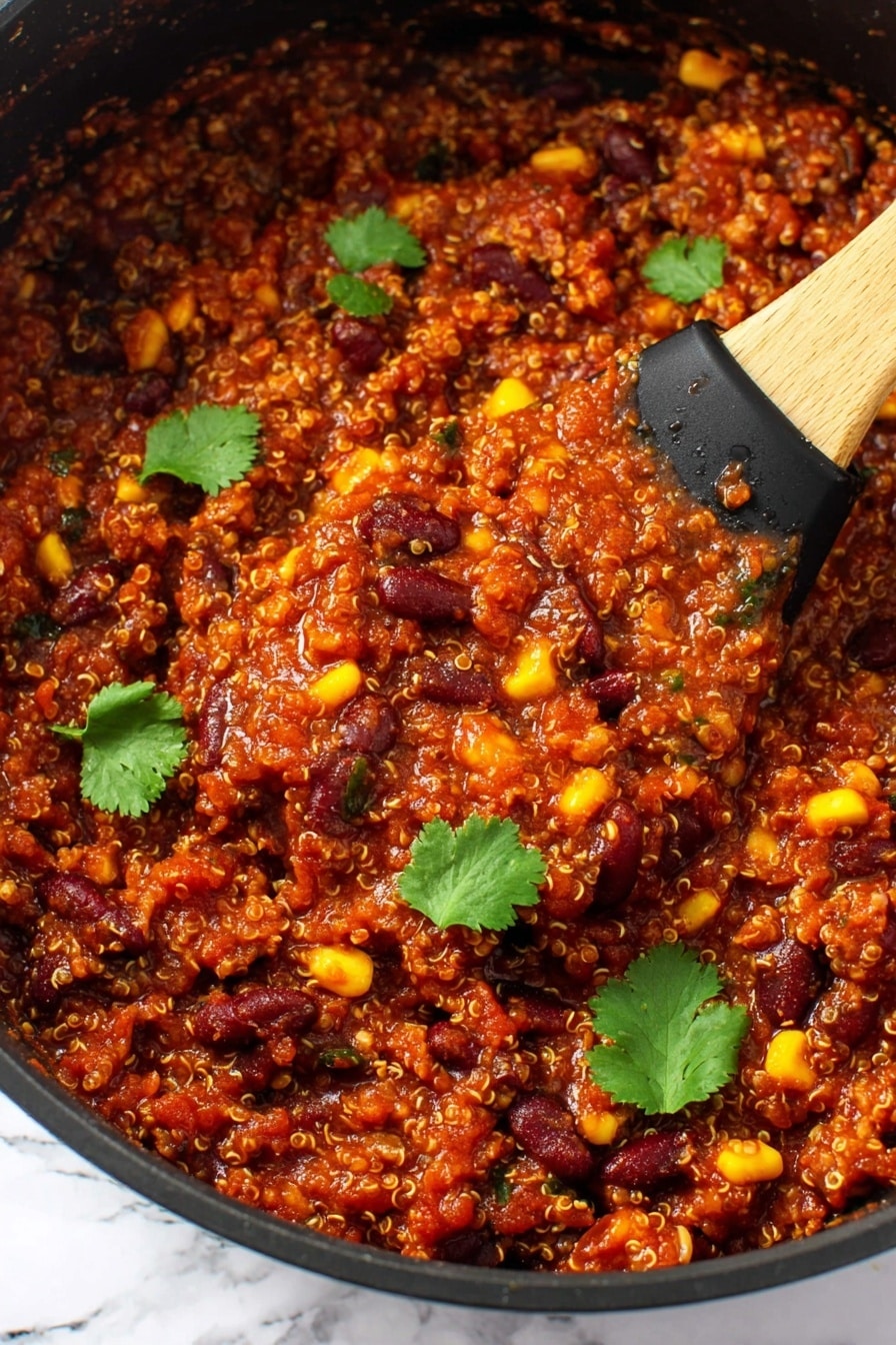 The image shows a thick, rich mixture in a black pan filled with three main layers: grains that look like tiny beige quinoa, dark red kidney beans, and bright yellow corn kernels, all mixed with a deep red tomato-based sauce. The texture is chunky with small bits of vegetables and beans visible throughout. There are several fresh green cilantro leaves scattered on top for contrast. A wooden spoon with a black silicone head is scooping some of the mixture from the pan. The background is a white marbled texture. Photo taken with an iphone --ar 2:3 --v 7 - Slow Cooker Vegan Bean & Quinoa Chili, vegan chili recipe, healthy vegan chili, hearty plant-based chili, easy vegan dinner