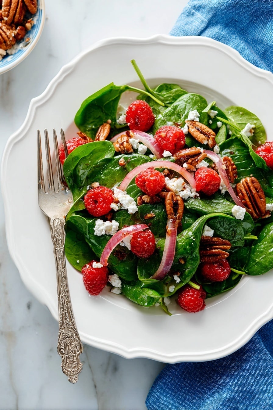 A white scalloped plate holds a fresh salad with three main layers: the base layer has bright green spinach leaves with soft texture, the middle layer has thin slices of pale pink-red onions scattered evenly, and the top layer features bright red raspberries, crumbled white cheese, and brown pecan halves arranged to create color contrast. A silver fork with an ornate handle rests on the left side of the plate, partially on the salad. The plate is placed on a white marbled surface with a blue cloth napkin nearby. Photo taken with an iphone --ar 2:3 --v 7 - Spinach Salad with Goat Cheese, Raspberries, and Toasted Pecans,healthy raspberry spinach salad,easy gourmet salads,fresh spinach salad recipes,nutty berry salad