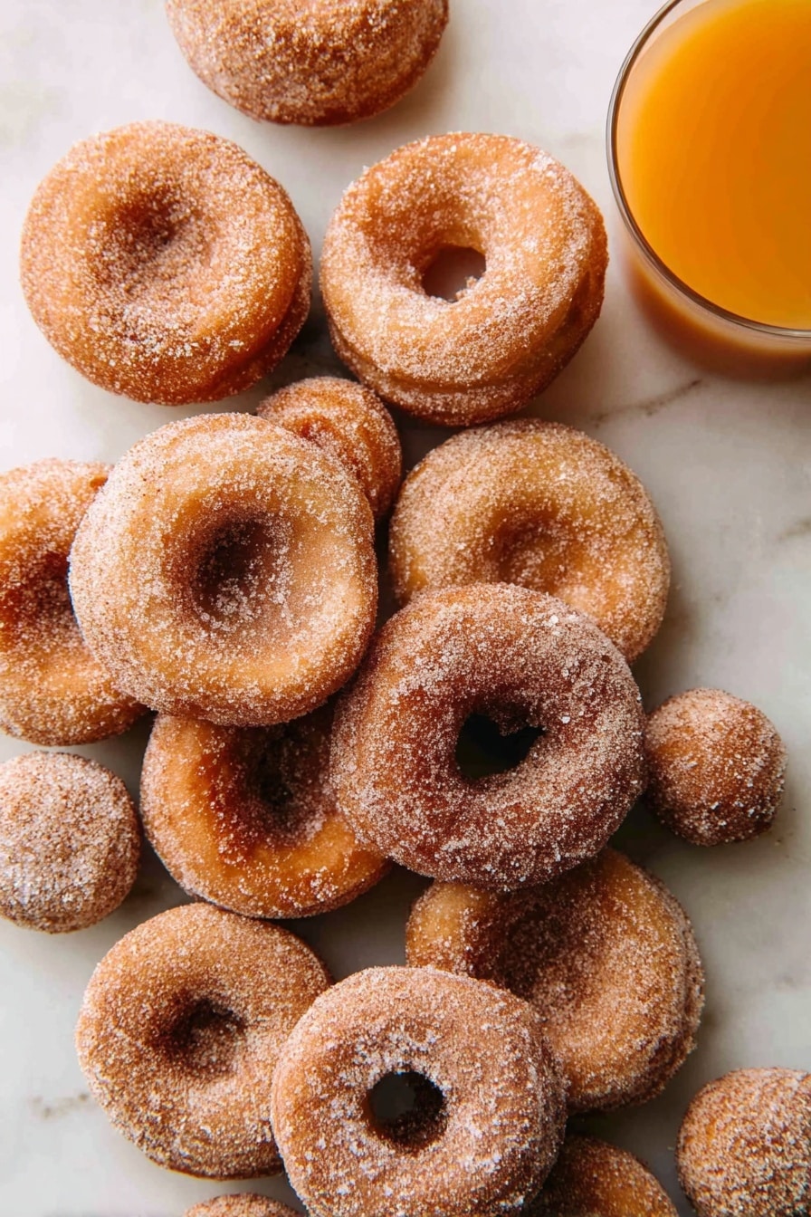 A pile of about a dozen small round donuts with light brown color, coated evenly with granulated sugar and cinnamon, arranged casually on a white marbled surface; two small round donut holes are also visible among the donuts, and at the top right corner, a clear glass of orange-colored juice is partially shown. photo taken with an iphone --ar 2:3 --v 7 - Apple Cider Donuts, Apple Cider Donuts Recipe, Fall Donuts, Homemade Apple Cider Donuts, Spiced Donut Recipe