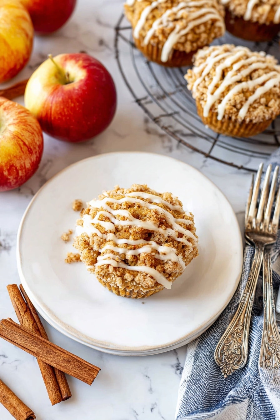 A single round muffin sits in the middle of a white plate, topped with a crumbly golden-brown streusel layer and drizzled with thick white icing in uneven stripes. The plate is on a white marbled surface surrounded by three shiny red and yellow apples, two cinnamon sticks, and a blue and white striped cloth. To the right, a wire cooling rack holds several more muffins with the same crumb topping and white icing drizzle. Two ornate silver forks rest beside the plate. The scene is bright and cozy, with warm autumn colors and textures. photo taken with an iphone --ar 2:3 --v 7 - Cinnamon Apple Muffins with Crumble Topping and Glaze, apple muffin recipes, fall breakfast muffins, easy apple muffins, cozy baking recipes