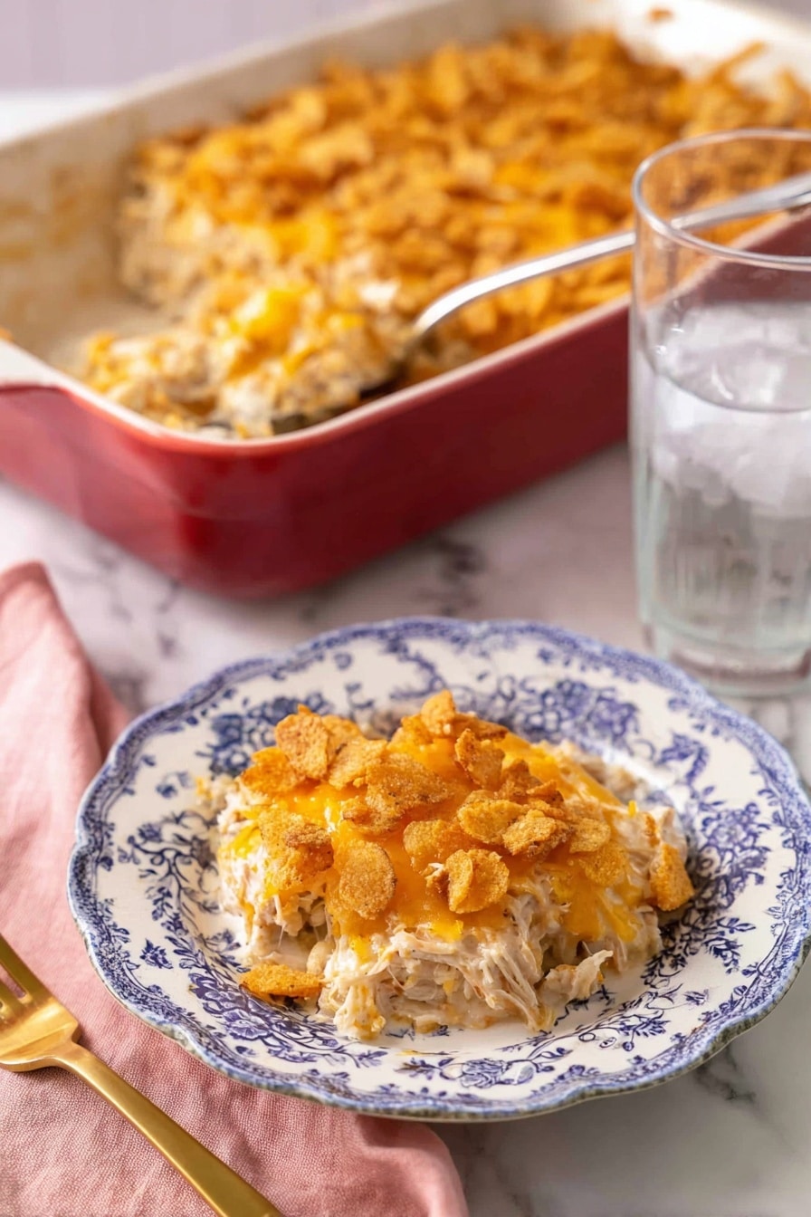 A portion of a creamy casserole is served on a white plate with blue floral patterns, showing three main layers: the bottom layer is shredded white meat mixed with a light creamy sauce, the middle layer has melted orange cheese blended with the meat, and the top layer consists of crushed crispy crackers with more melted cheese giving a golden-brown, crunchy texture. Behind the plate, there is a large red baking dish filled with the same casserole, with a silver serving spoon inside. To the right of the plate is a clear glass of ice water, and to the left is a gold fork resting on a pink cloth napkin, all placed on a white marbled surface. photo taken with an iphone --ar 2:3 --v 7 - Creamy Chicken Casserole, easy chicken casserole, comforting chicken dinner, cheesy chicken bake, family-friendly casserole