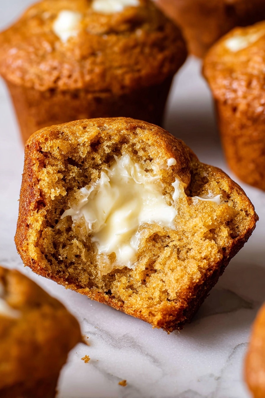 The image shows a close-up of a muffin broken in half, revealing a soft, moist texture inside with a spread of creamy, melted butter on one side. The muffin’s outer crust is rich golden brown and slightly crisp, while the inside is a dense, honey-brown color with small air pockets. Surrounding the main muffin piece, there are other muffins blurred slightly in the background, all with a similar warm golden outer color. The surface beneath the muffins is a white marbled texture. photo taken with an iphone --ar 2:3 --v 7 - Perfect Pumpkin Muffins, pumpkin muffins recipe, fall muffin recipes, moist pumpkin muffins, cozy spice muffins