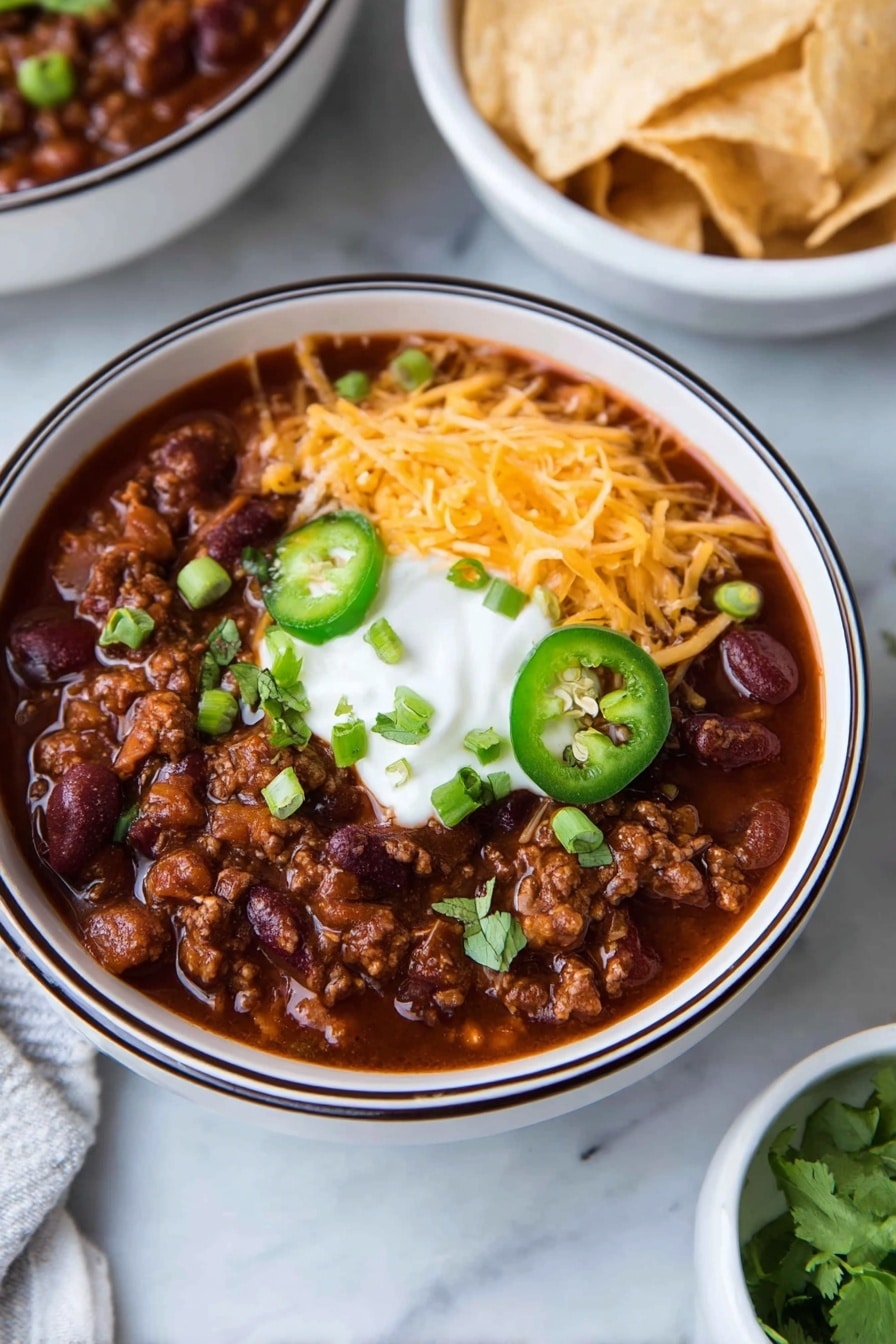 A white bowl with a dark rim holds a thick dark red chili filled with beans and ground meat. On top, there is a layer of shredded orange cheddar cheese on one side, a dollop of white sour cream in the center, sliced green jalapeño rings below, and chopped green onions and cilantro scattered around. In the background, there is a white bowl filled with light-colored tortilla chips on a white marbled surface. Photo taken with an iphone --ar 2:3 --v 7 - Healthy Turkey Chili, healthy turkey chili recipe, easy turkey chili, low-calorie chili, protein-packed chili