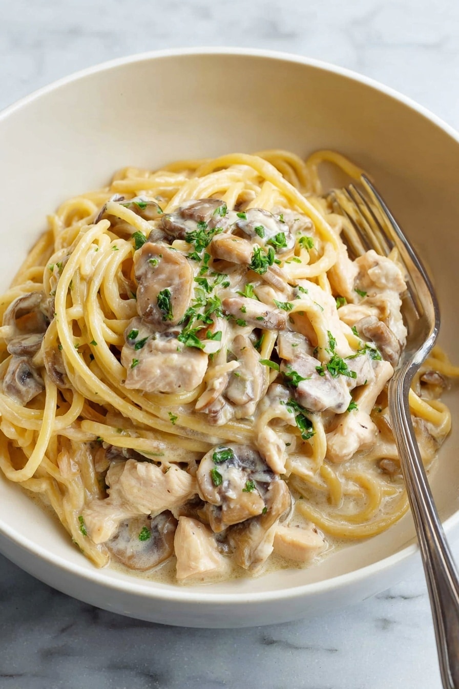 A white bowl holds creamy pasta with three main layers: at the bottom are light brown mushroom pieces coated in sauce, on top are thick, pale yellow spaghetti noodles mixed with tender white chicken pieces, all covered in a smooth, light cream sauce. Small green parsley bits are scattered on top, adding a touch of color. A silver fork rests inside the bowl on the right side. The bowl sits on a white marbled surface. Photo taken with an iphone --ar 2:3 --v 7 - Creamy Chicken Tetrazzini Bake, chicken tetrazzini bake, creamy pasta casserole, easy chicken baked pasta, comfort food recipes