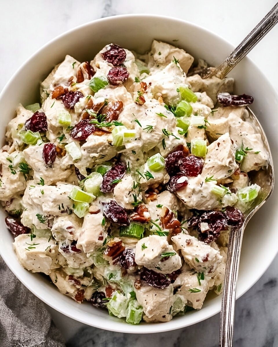 The image shows a white bowl filled with a creamy chicken salad. The salad has multiple layers: light beige chunks of cooked chicken, bright green pieces of celery and chopped green onions, and dark red dried cranberries spread throughout. Small pieces of brown pecans add texture, and the salad is mixed with a white creamy dressing that coats all the ingredients gently. Small green herb pieces are sprinkled on top, enhancing the texture and color variety. A silver spoon rests inside the bowl, ready to serve, all placed on a white marbled surface. photo taken with an iphone --ar 4:5 --v 7