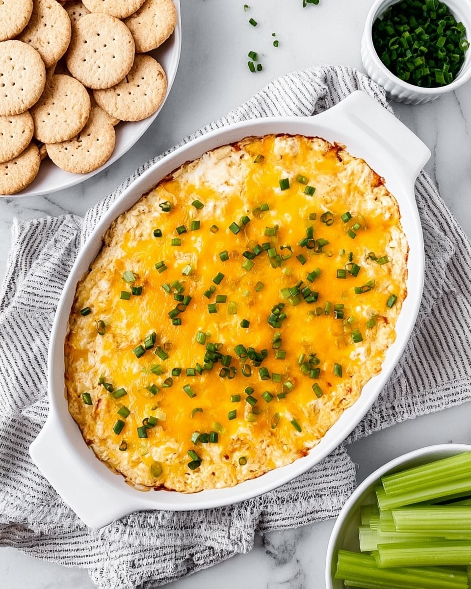 A white oval dish filled with a creamy baked dip that has a melted golden-yellow cheese layer on top, sprinkled with small green chive pieces evenly spread over the surface. The dish is set on a white and black striped cloth on a white marbled background. Around the dish, there are light beige crackers on the left side and green celery sticks in a white bowl at the bottom. At the top right, there is a small white bowl with extra chopped chives. Photo taken with an iphone --ar 4:5 --v 7