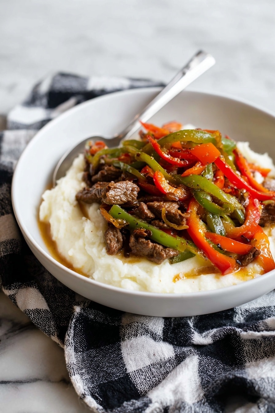 A white bowl holds a dish with two main layers: the bottom layer is a smooth, creamy white mashed texture, slightly spread around the edges. On top, there is a colorful mix of thin strips of cooked green, red, and orange bell peppers, mixed with small pieces of brown cooked meat, all glistening with a light sauce. A silver spoon rests inside the bowl, angled towards the back. The bowl sits on a black and white checkered cloth, placed on a white marbled surface. photo taken with an iphone --ar 2:3 --v 7 - Pepper Steak with Onion, Pepper Steak with Onion recipe, savory pepper steak, quick beef stir-fry, flavorful flank steak with peppers