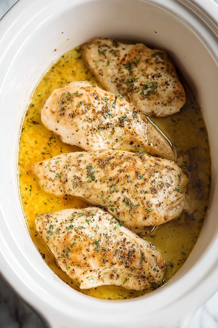 The image shows four pieces of cooked chicken placed side by side inside a white slow cooker. Each piece is light golden brown with some darker brown spots from cooking and sprinkled with green herbs and black pepper. The chicken sits in a shallow puddle of yellowish oil or broth that gives a slight shine on the surface. The inside of the slow cooker is white and smooth, contrasting with the warm tones of the chicken and liquid. The setting features a white marbled background. photo taken with an iphone --ar 2:3 --v 7 - Slow Cooker Juicy & Seasoned Chicken Breast, tender slow cooker chicken, easy slow cooker chicken recipes, moist chicken breast slow cooker, flavorful chicken dinner