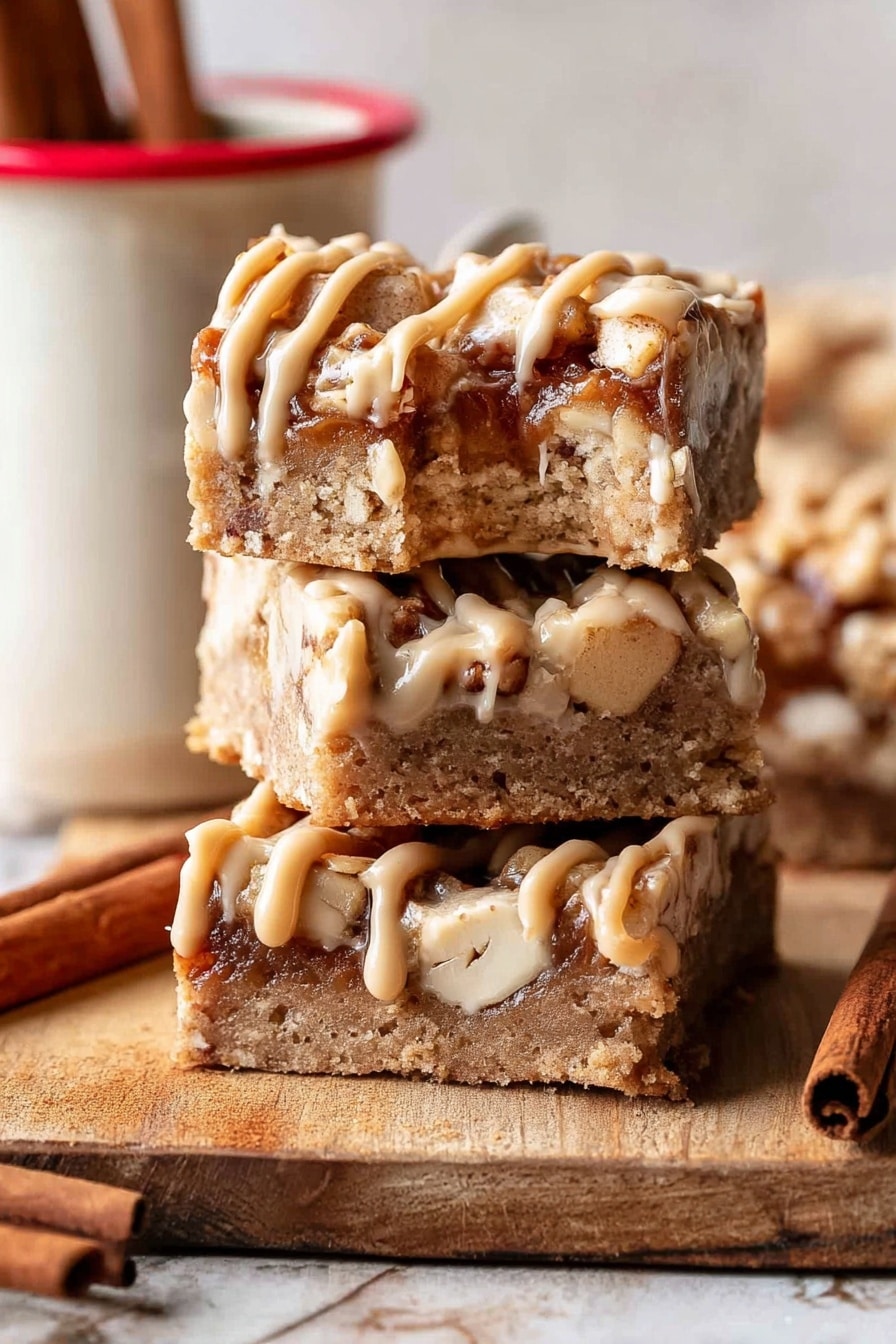 The image shows three square bars stacked vertically on a wooden surface with a single bite taken from the top bar. Each bar has a thick, crumbly base layer in light brown with small white nut pieces embedded inside. On top of the base, there is a layer of darker brown chunks with a soft texture, drizzled with thick, pale caramel-colored frosting in thin, uneven lines. A cinnamon stick is placed near the bottom left corner, and a white mug with a red rim and two cinnamon sticks inside is partially visible to the right. The background is a white marbled texture. Photo taken with an iphone --ar 2:3 --v 7 - Brown Butter Apple Blondies with Cinnamon Maple Glaze, fall dessert recipes, cozy apple blondies, homemade blondie recipes, apple cinnamon dessert