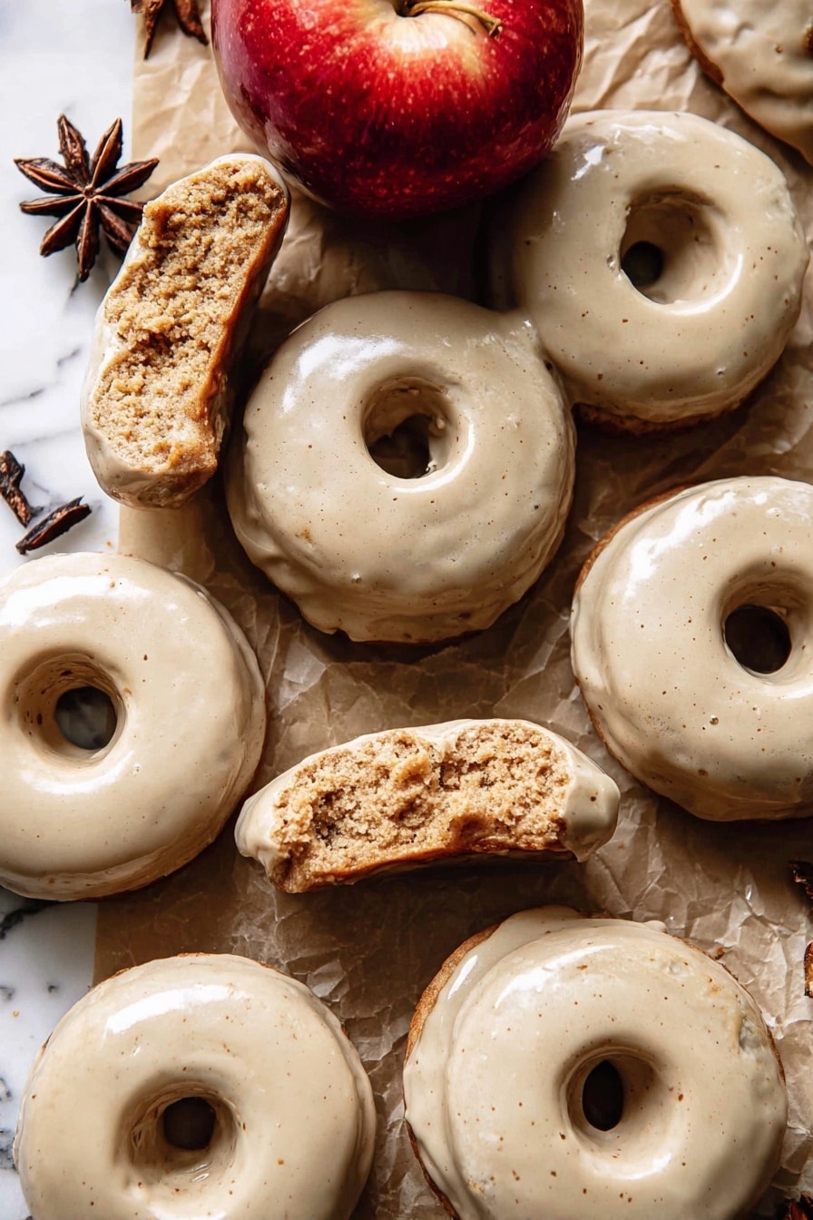 The image shows a group of doughnuts with a smooth, light beige glaze covering their top surfaces. Each doughnut has one round hole in the center and a slightly rough, crumbly texture inside where some are broken or bitten. The doughnuts are laid on a brown paper surface with pieces of white parchment paper scattered around. There is a shiny red apple near the center top of the image and a dark brown star anise near the upper left side. The background is a white marbled texture. photo taken with an iphone --ar 2:3 --v 7 - Baked Apple Cider Doughnuts with Cinnamon Maple Glaze, fall apple cider doughnuts, baked apple cinnamon doughnuts, easy seasonal apple donuts, cozy autumn baked treats