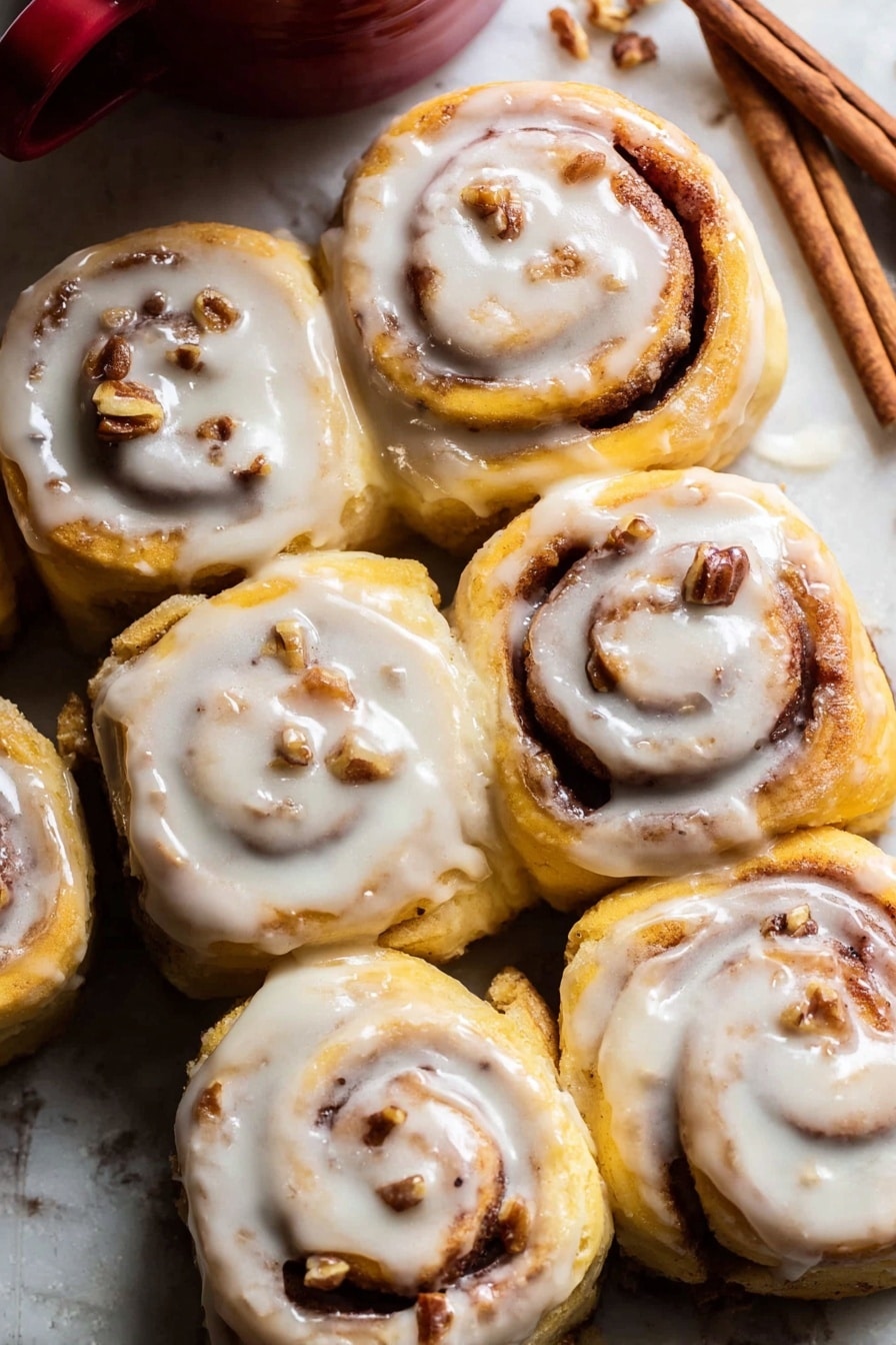 A close-up view of six cinnamon rolls arranged closely together on a white marbled surface. Each roll shows a soft golden-yellow dough spiraled with a dark cinnamon and sugar filling, with small pieces of nuts embedded within. A creamy white icing with a smooth texture generously covers the top of each cinnamon roll, slightly dripping down the sides. In the upper background, a few cinnamon sticks lay on the white marbled surface near the top right corner, next to a red ceramic cup partially visible. The warm tones of the rolls contrast nicely with the white icing and the white marbled surface, giving a fresh and inviting look. photo taken with an iphone --ar 2:3 --v 7 - Apple Cinnamon Rolls with Brown Butter Maple Icing, cinnamon rolls with apple and brown butter, homemade apple cinnamon rolls, easy apple cinnamon roll recipe, cozy breakfast cinnamon rolls