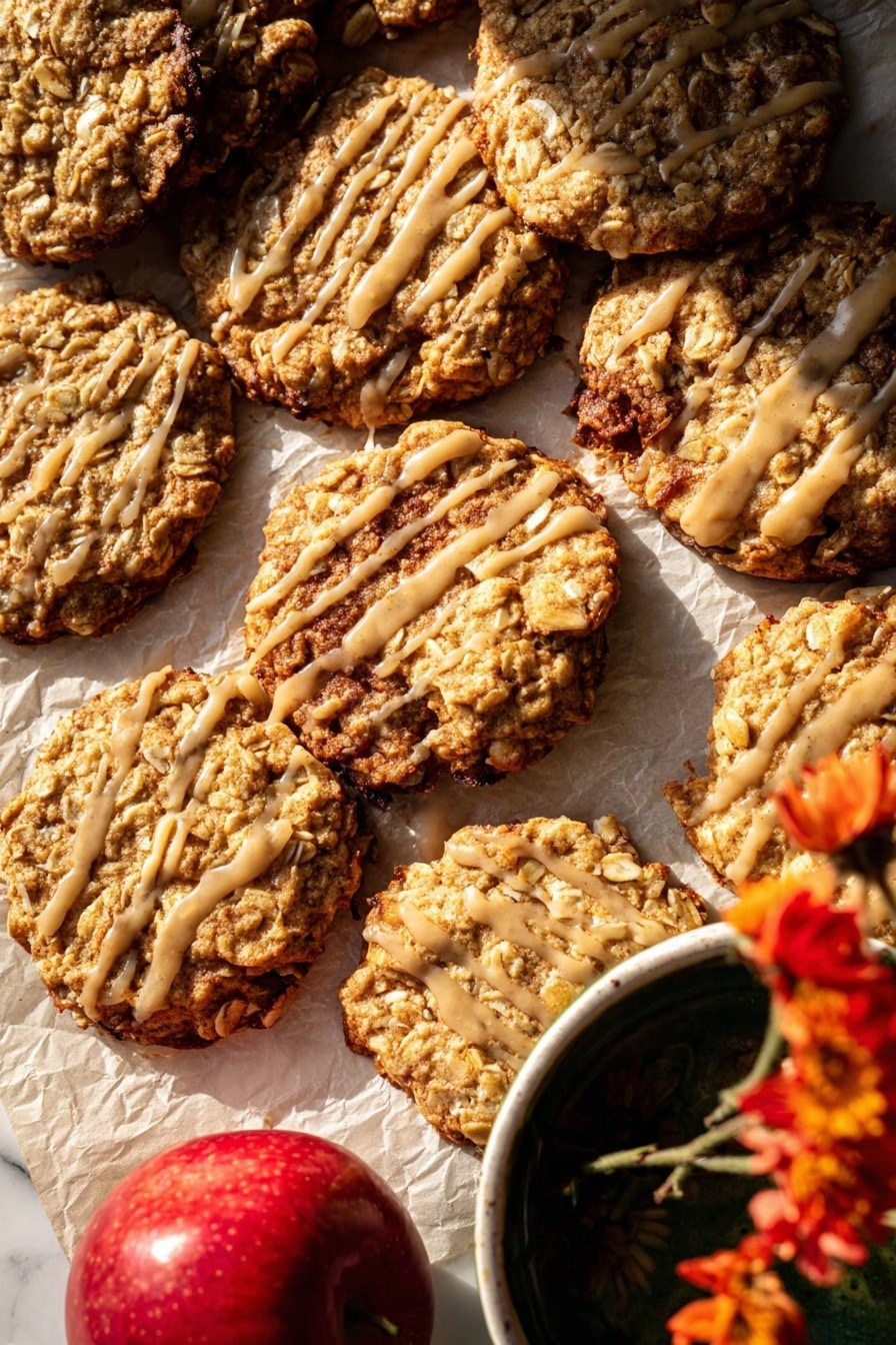 The image shows a close-up of several oatmeal cookies with a slightly rough texture from oats and nuts, arranged unevenly on parchment paper over a white marbled surface. Some cookies are stacked, while others lay flat, revealing their golden-brown baked edges with a few darker spots. A few cookies are drizzled with a light beige glaze in thin, irregular lines, adding shine and contrast to the surface. A bright red apple is partially visible in the bottom left, and the corner of a white bowl with a dark liquid and a yellow flower is seen in the top right, adding color variety. The warm lighting highlights the cookies' texture and the glaze’s glossiness. photo taken with an iphone --ar 2:3 --v 7 - Iced Apple Cider Caramel Oatmeal Cookies, apple cider oatmeal cookies, caramel oatmeal cookies, fall cookies with apple cider, chewy caramel oatmeal treats