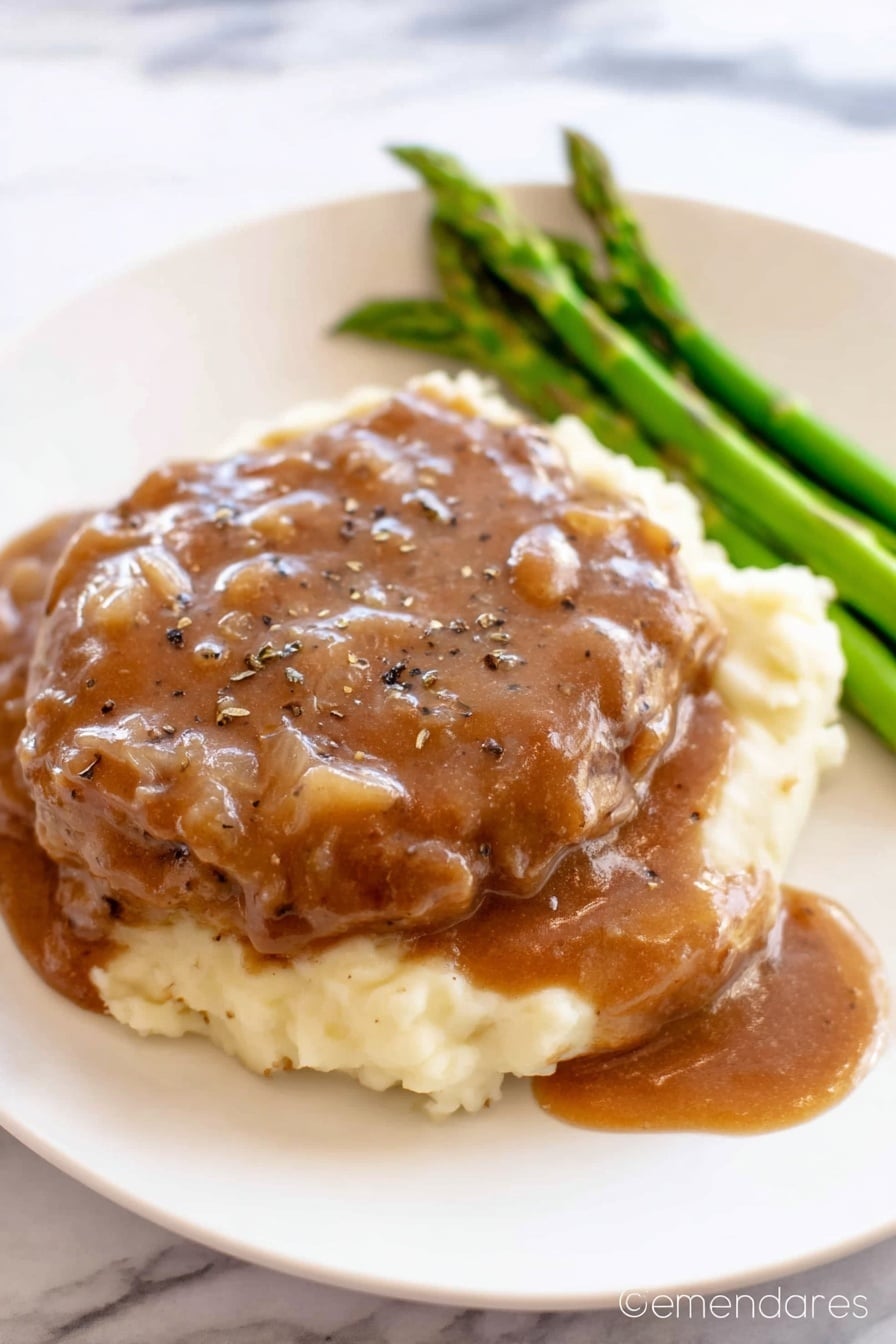 The image shows a white plate with three layers of food. The bottom layer is creamy mashed potatoes, which look soft and smooth in a pale cream color. On top of the mashed potatoes is a thick piece of cooked meat covered generously with a brown gravy sauce that has small onion bits and black pepper sprinkled on top. To the side of the meat, there is a small bunch of cooked green asparagus spears that add a fresh green color. The white plate sits on a white marbled surface. Photo taken with an iphone --ar 2:3 --v 7 - Crock Pot Cube Steak, slow cooker cube steak recipe, easy cube steak dinner, tender beef stew, hearty slow cooker meals