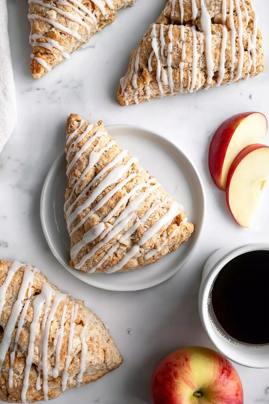 Several triangular scones are arranged on a white marbled surface, each covered in a light brown, crumbly texture with white icing drizzled in even stripes across the top. One scone sits on a simple white plate centered in the image, accompanied by two slices of red and yellow apple on the right side of the plate. Nearby, a whole apple with red and yellow skin is partly visible at the bottom right. In the upper right corner, a white cup filled with black coffee adds contrast. The scene is bright and clean, highlighting the soft texture of the scones and fresh fruit. photo taken with an iphone --ar 2:3 --v 7 - Easy Apple Scones, apple scones recipe, flaky scones with apple, homemade apple scones, fall breakfast ideas