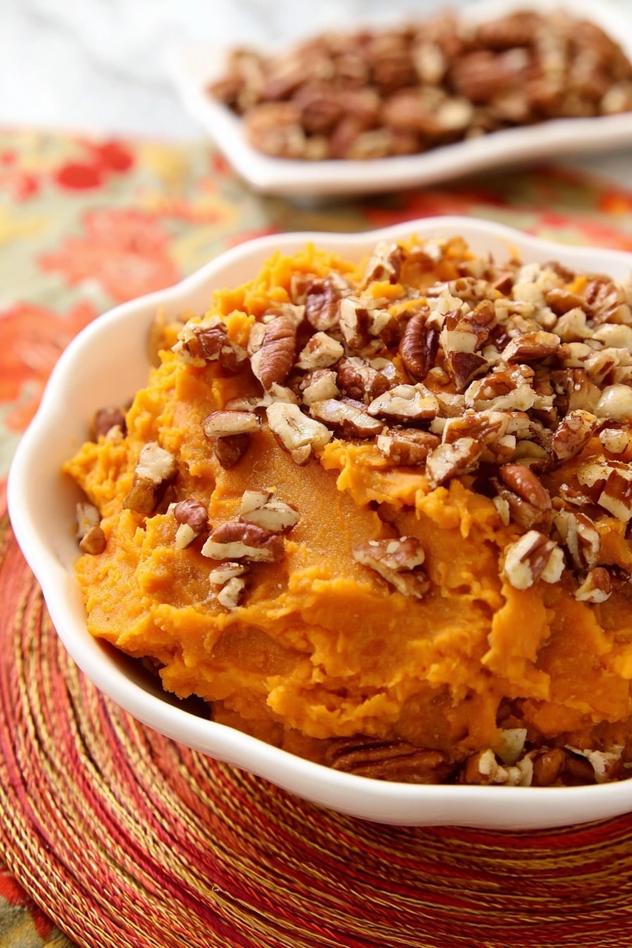 A white scalloped bowl holds a single layer of smooth, bright orange mashed sweet potatoes topped generously with a layer of chopped pecans, which add a textured, brown, and crunchy contrast. In the background, a white dish contains more pecan pieces, slightly out of focus. The bowl and pecans rest on a woven, multi-colored orange and red placemat, with a shiny silver spoon placed nearby. The scene is set on a white marbled surface. Photo taken with an iphone --ar 2:3 --v 7 - Slow Cooker Sweet Potato Casserole with Toasted Pecans, sweet potato casserole, slow cooker sweet potato side dish, holiday sweet potato recipes, easy slow cooker side dish